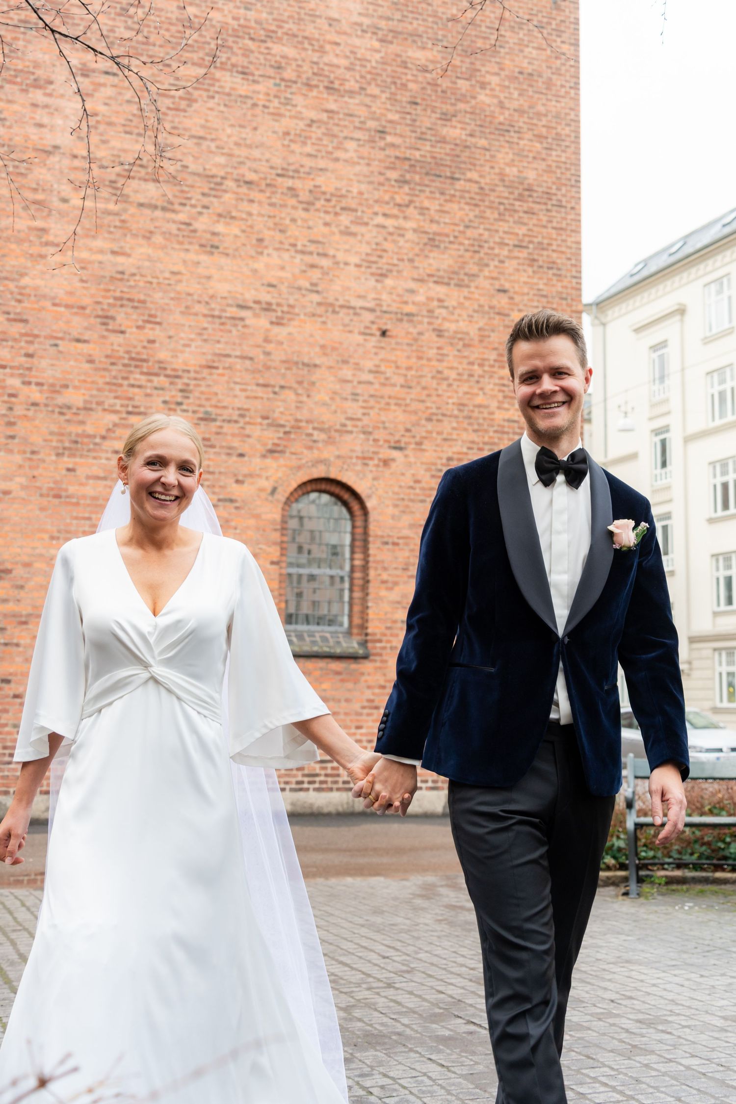 A happy couple in formal attire, a bride in a white dress and a groom in a tuxedo, walking hand-in-hand on a cobblestone street.