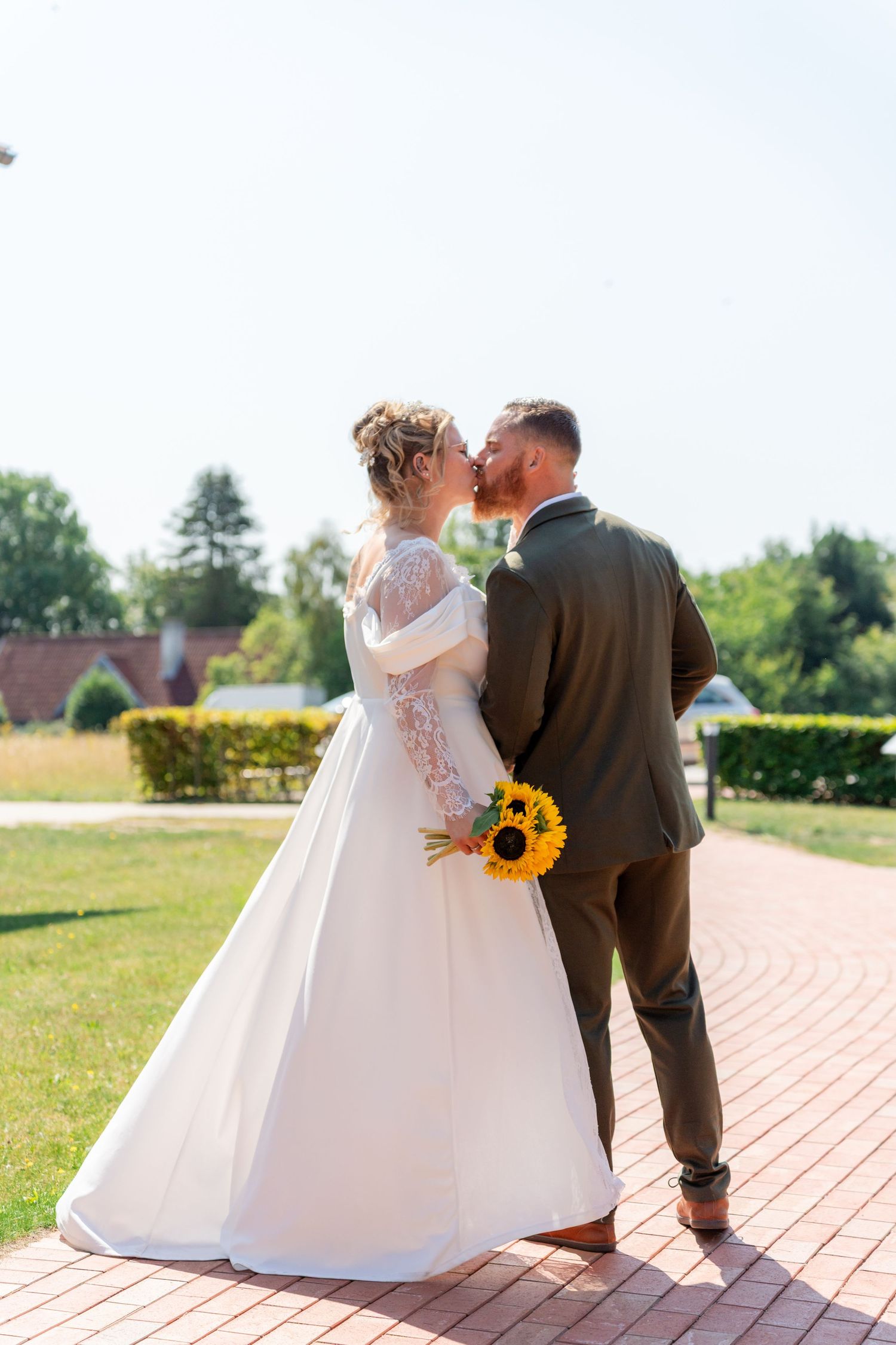 Bride and groom sharing a kiss outdoors, bride holding sunflower bouquet in white lace gown, groom in dark suit.