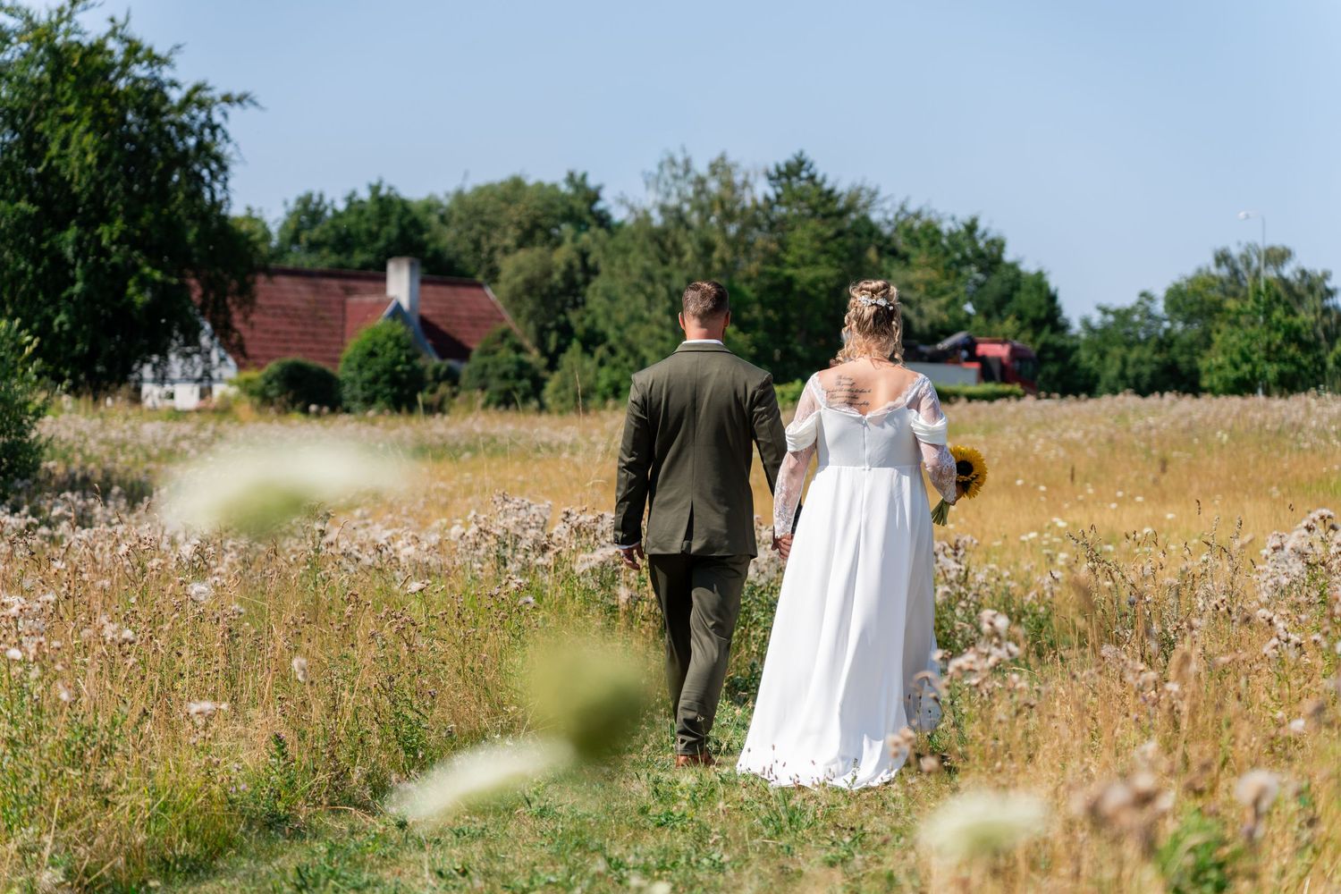 Bride and groom walking hand in hand through a wildflower meadow with a rustic farmhouse in the background.