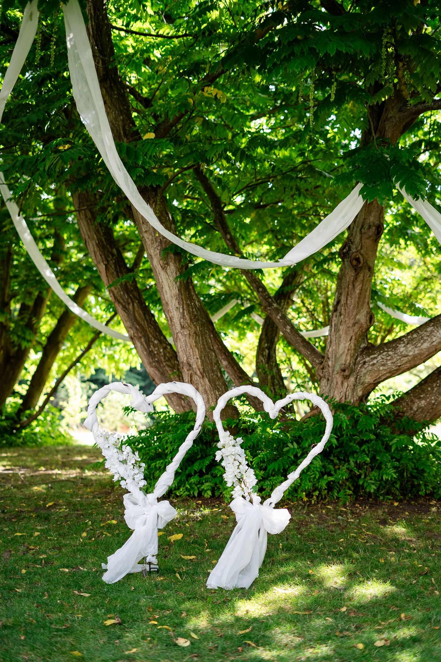Two white heart-shaped wedding decorations with floral accents stand on green grass beneath a tree draped with white ribbon.