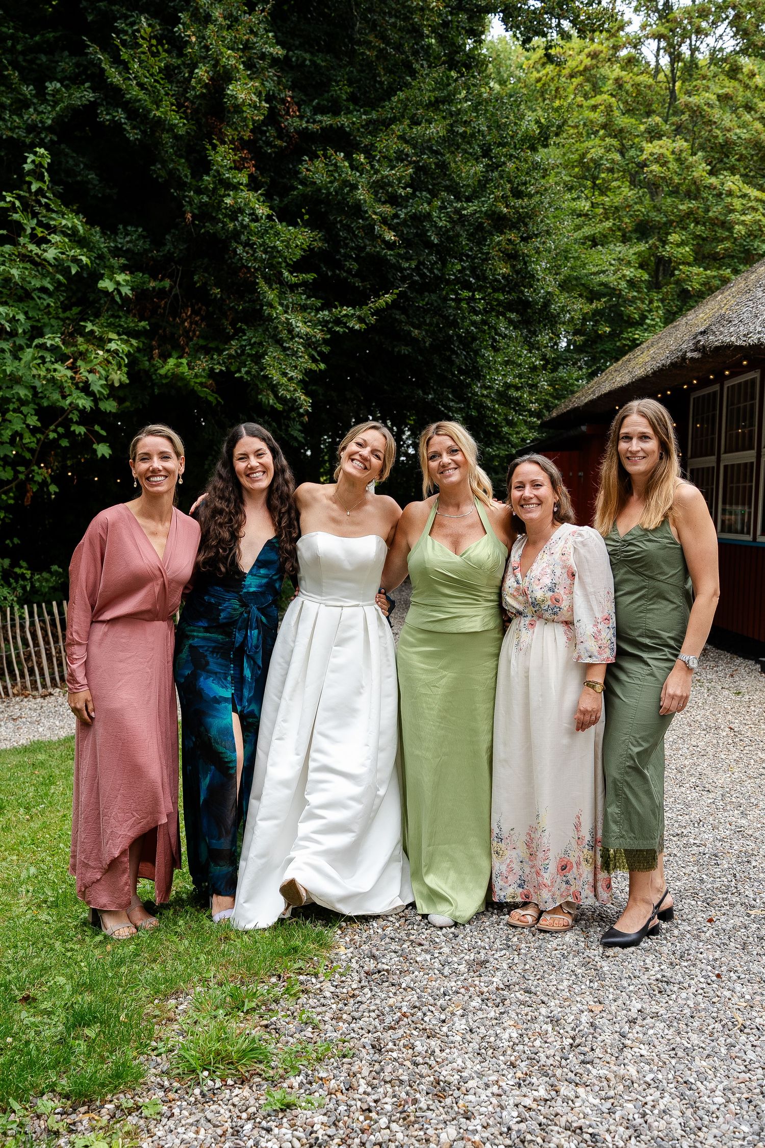 A bride in a white gown poses with five women in colorful dresses outdoors at a wedding celebration.