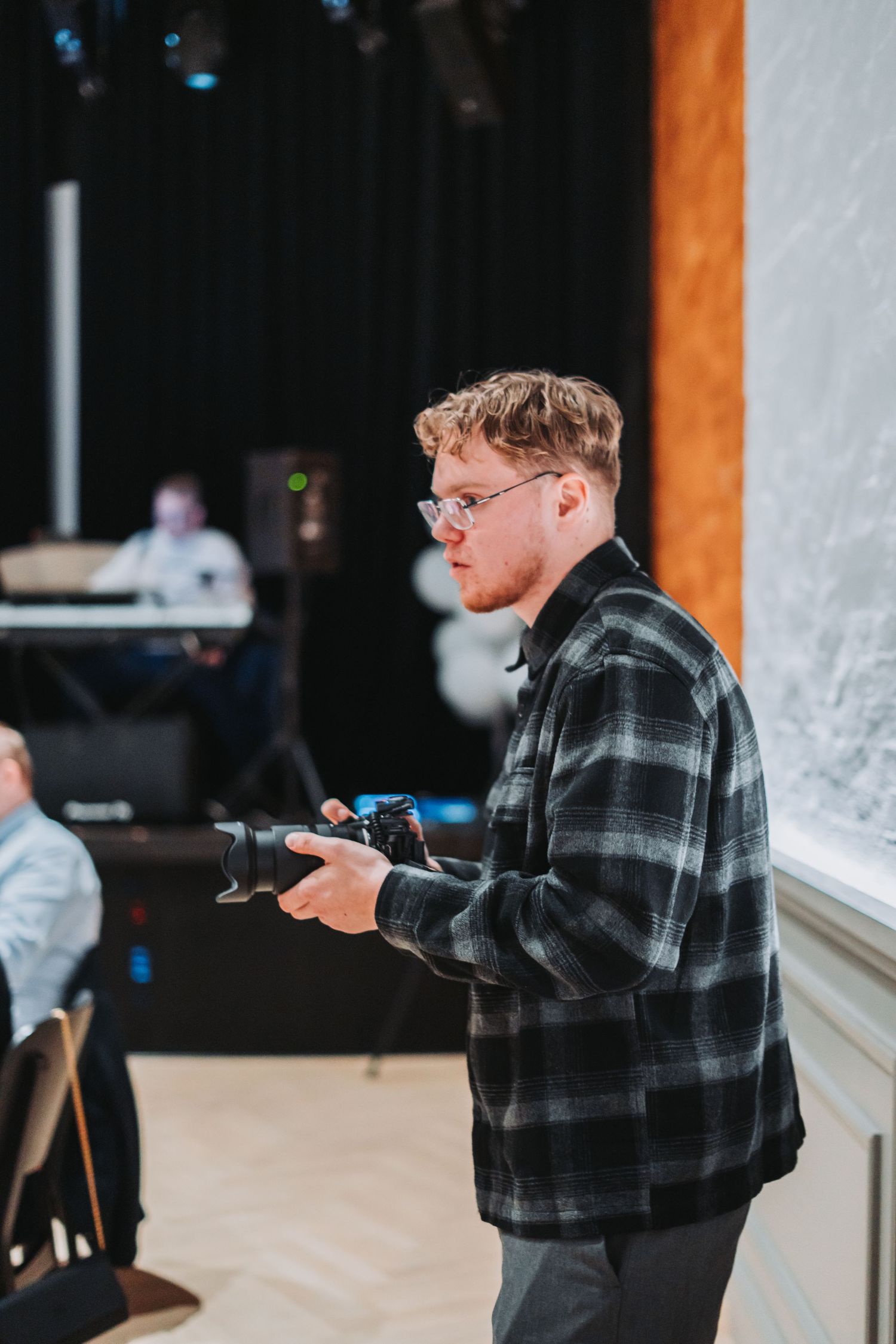 Photographer wearing plaid shirt checking camera settings in indoor venue.