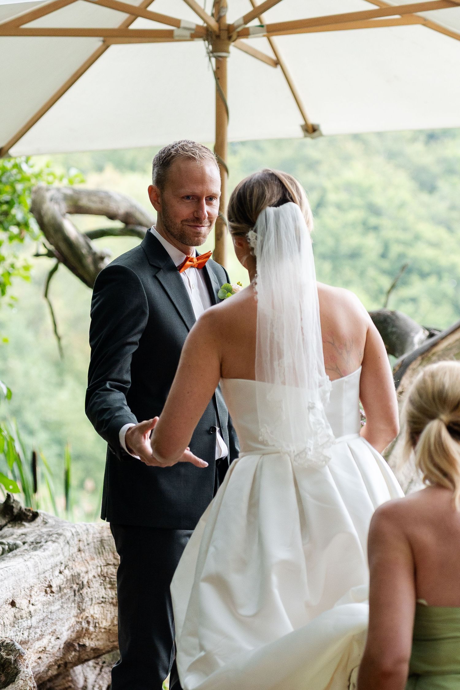 Groom in black suit with orange bow tie holds bride's hand during outdoor wedding ceremony under white umbrella.