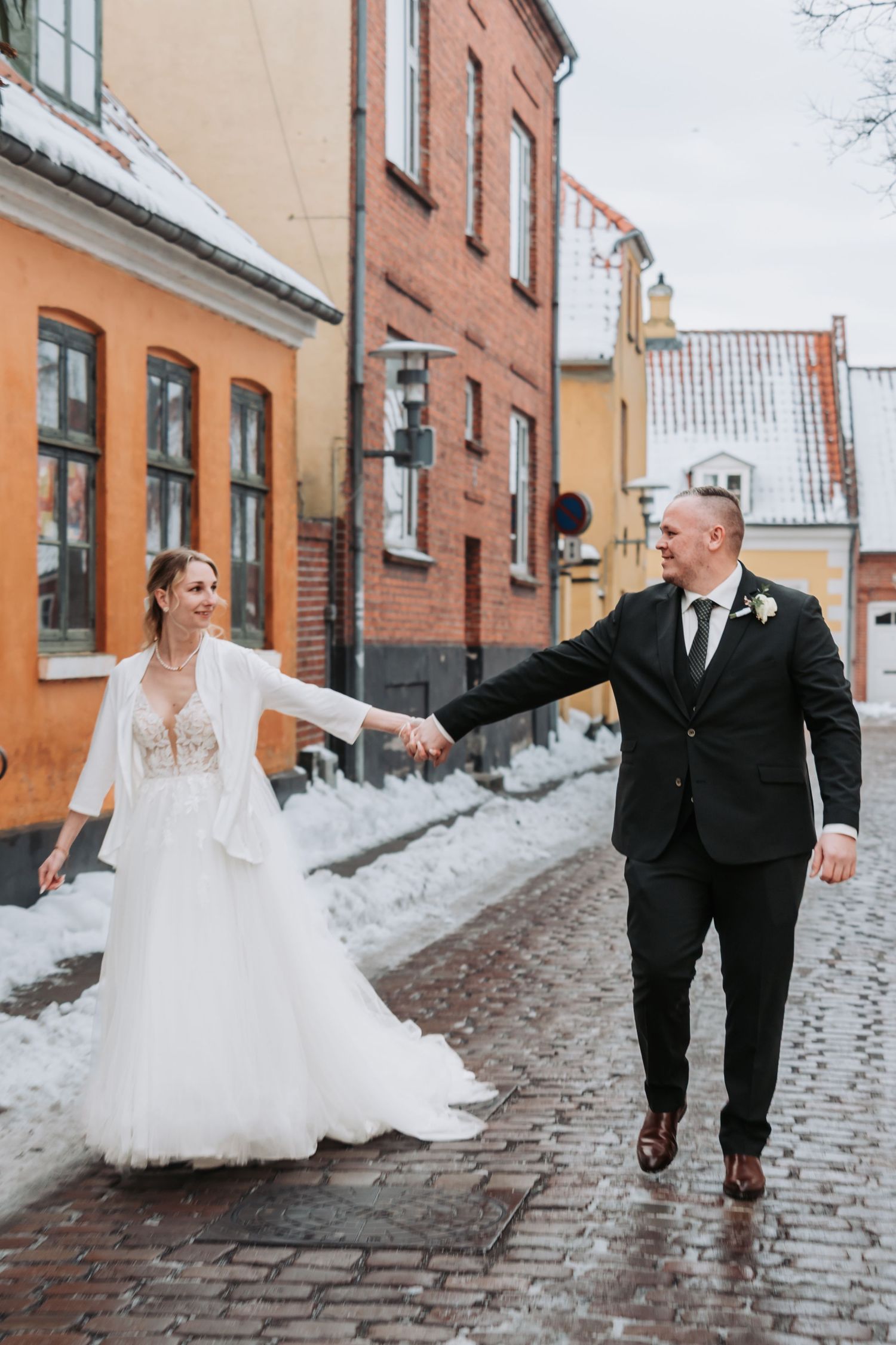 Wedding couple walking and twirling on snow-covered cobblestone street.