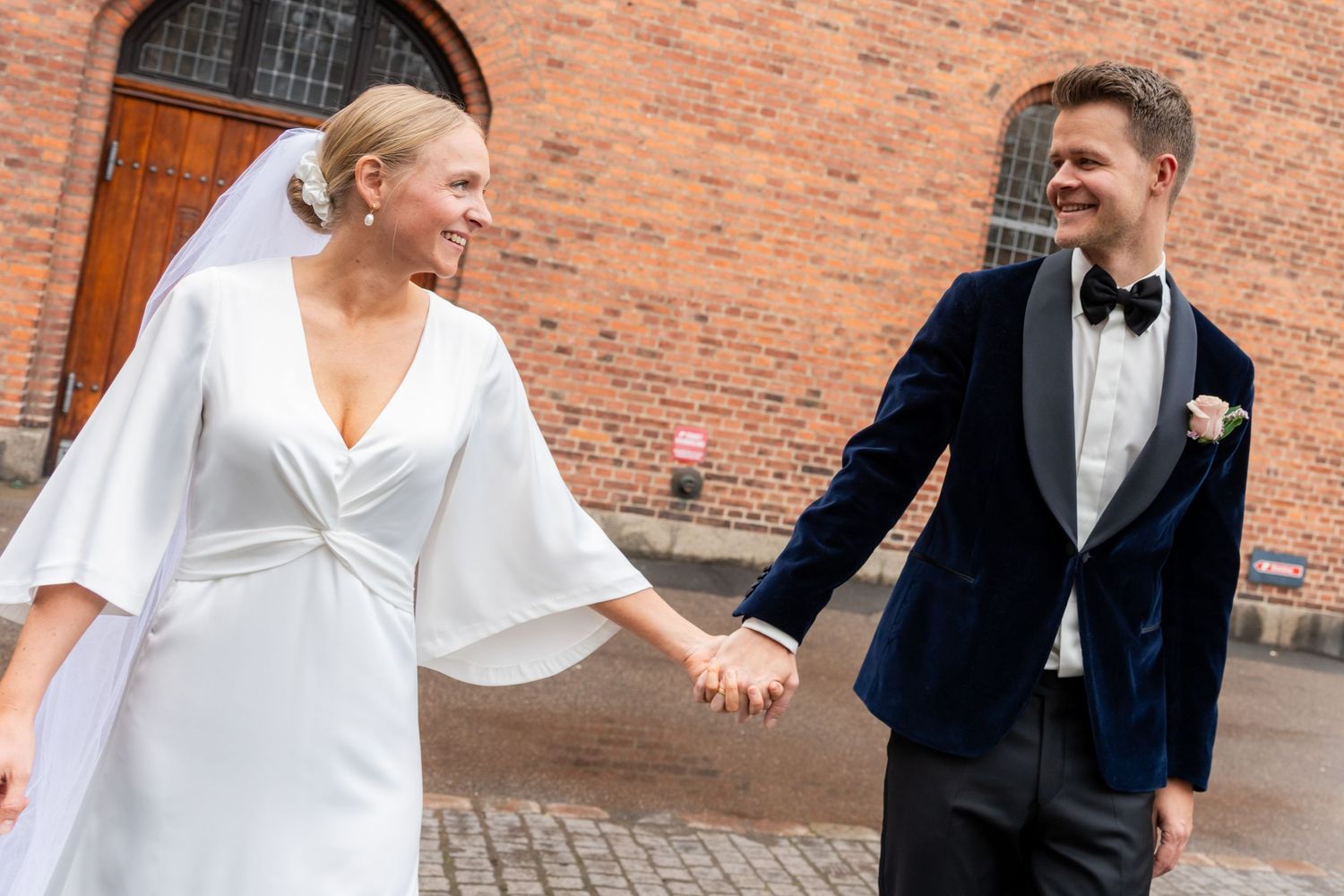 A bride in a white dress and groom in a tuxedo holding hands while standing in front of a brick building.