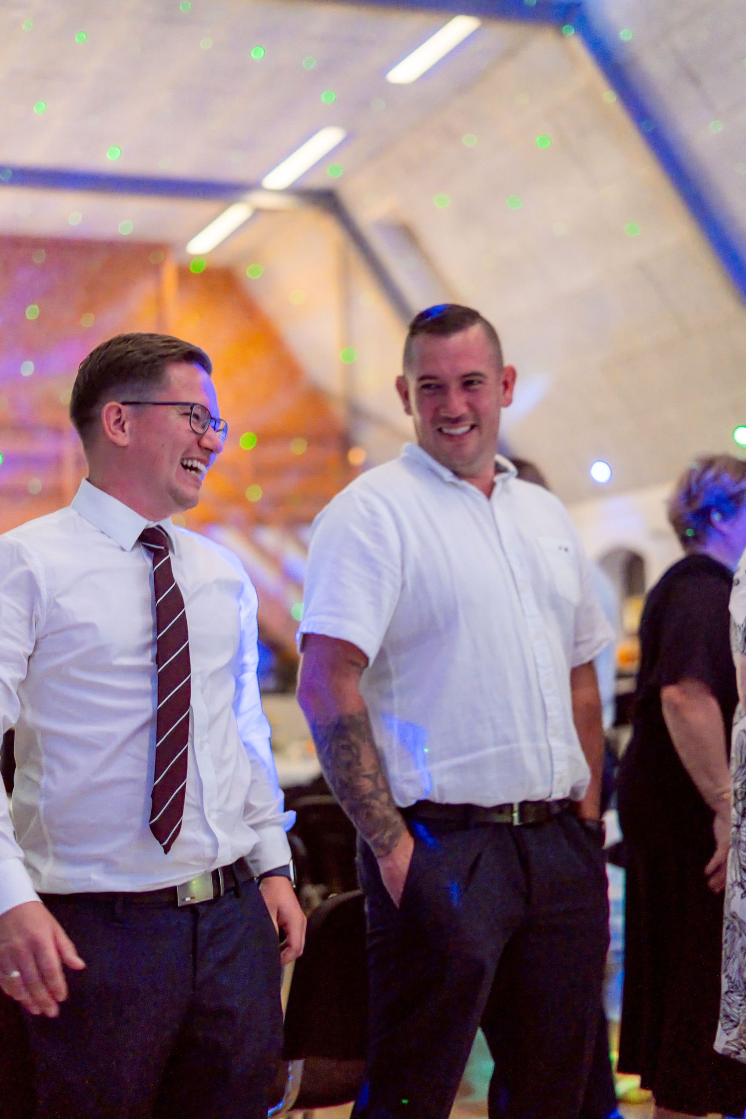Two men in white shirts laughing and socializing at a festive indoor event with colorful lights.