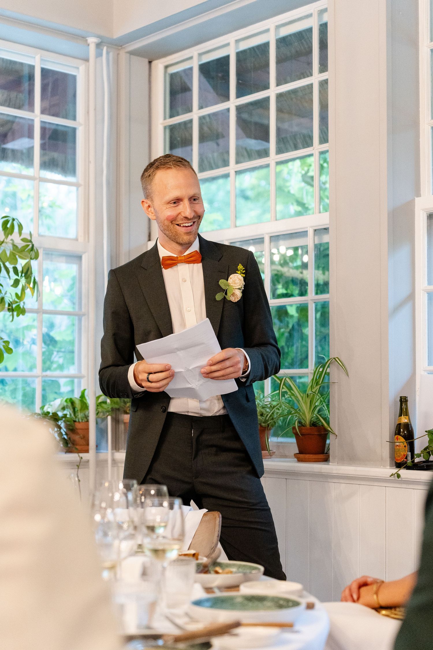 Smiling groom in black suit with orange bow tie and boutonniere holds speech notes at elegant wedding reception.
