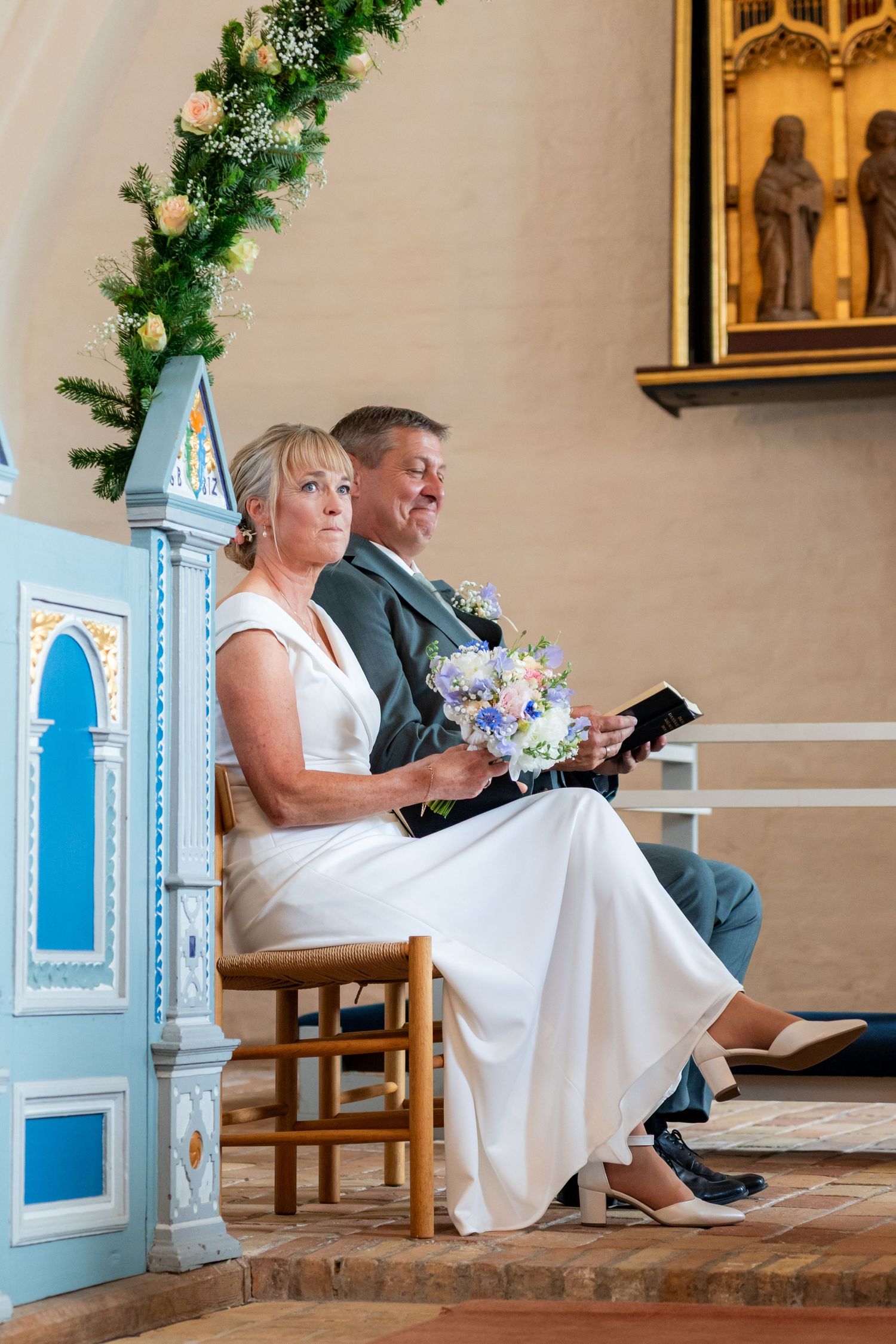 Bride and groom seated at church altar during wedding ceremony, bride holding colorful bouquet in white dress.