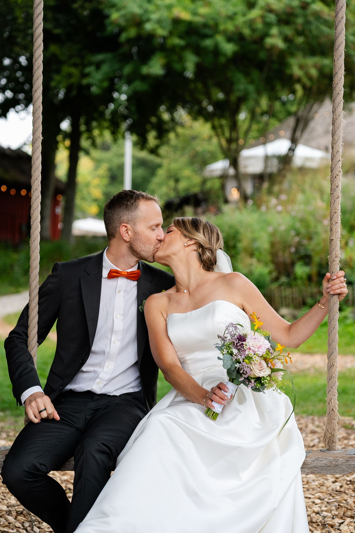 Bride and groom kissing on a rope swing outdoors, bride holding a bouquet, groom wearing black suit with orange bow tie.