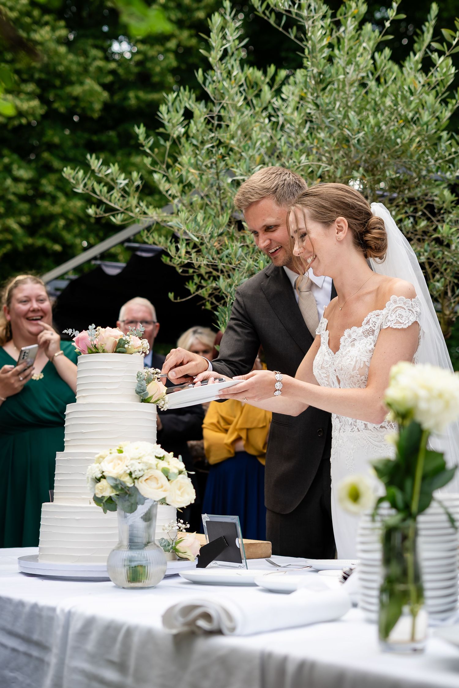 Bride and groom cutting white tiered wedding cake outdoors, smiling guests watching in background.