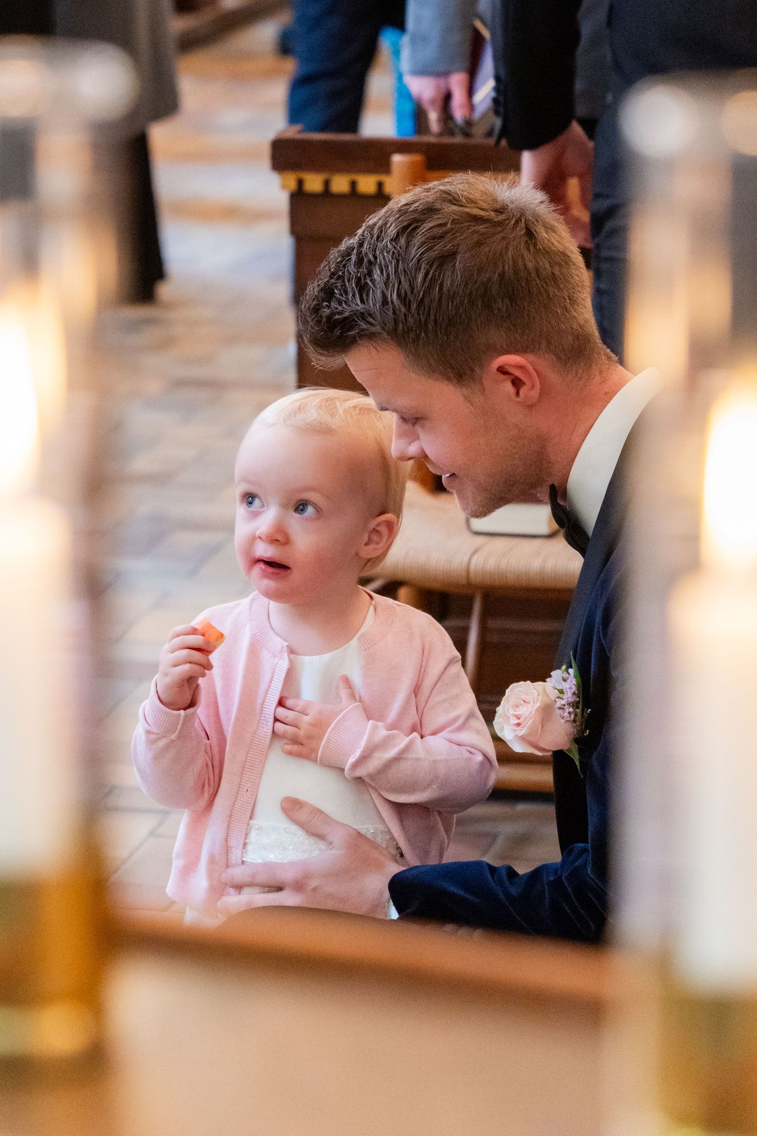 A man in a suit smiles at a baby girl in a pink cardigan during a wedding ceremony with candles in the foreground.