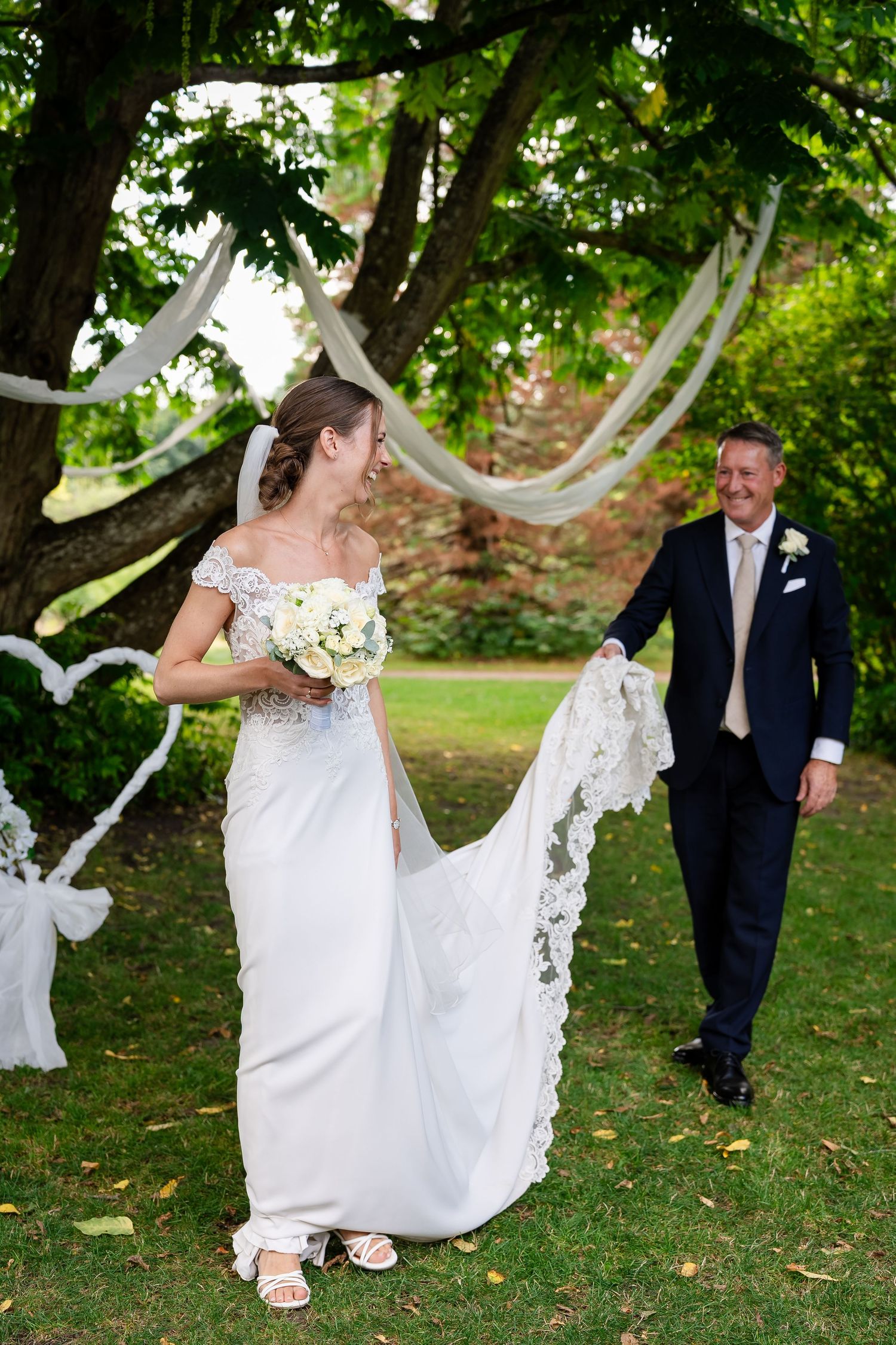 Bride in white lace gown holding bouquet walks with groom in navy suit during outdoor wedding ceremony.