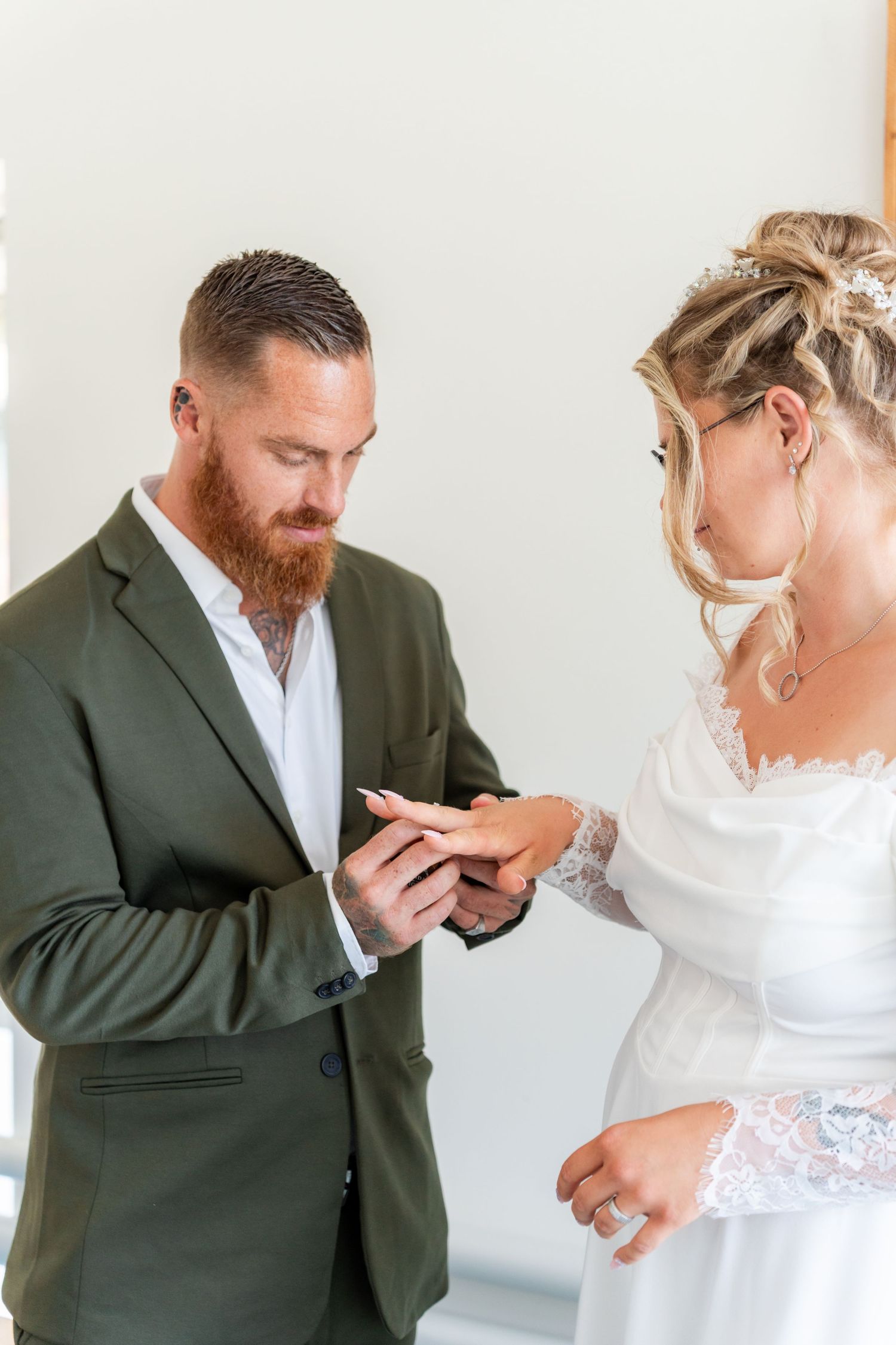Bride in white lace dress placing wedding ring on groom's finger, groom wearing green suit with red beard.