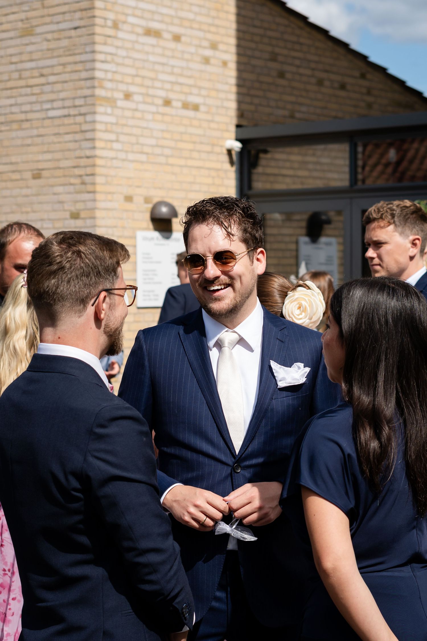 Smiling groom in navy pinstripe suit and sunglasses chatting with wedding guests outside a brick building on a sunny day.