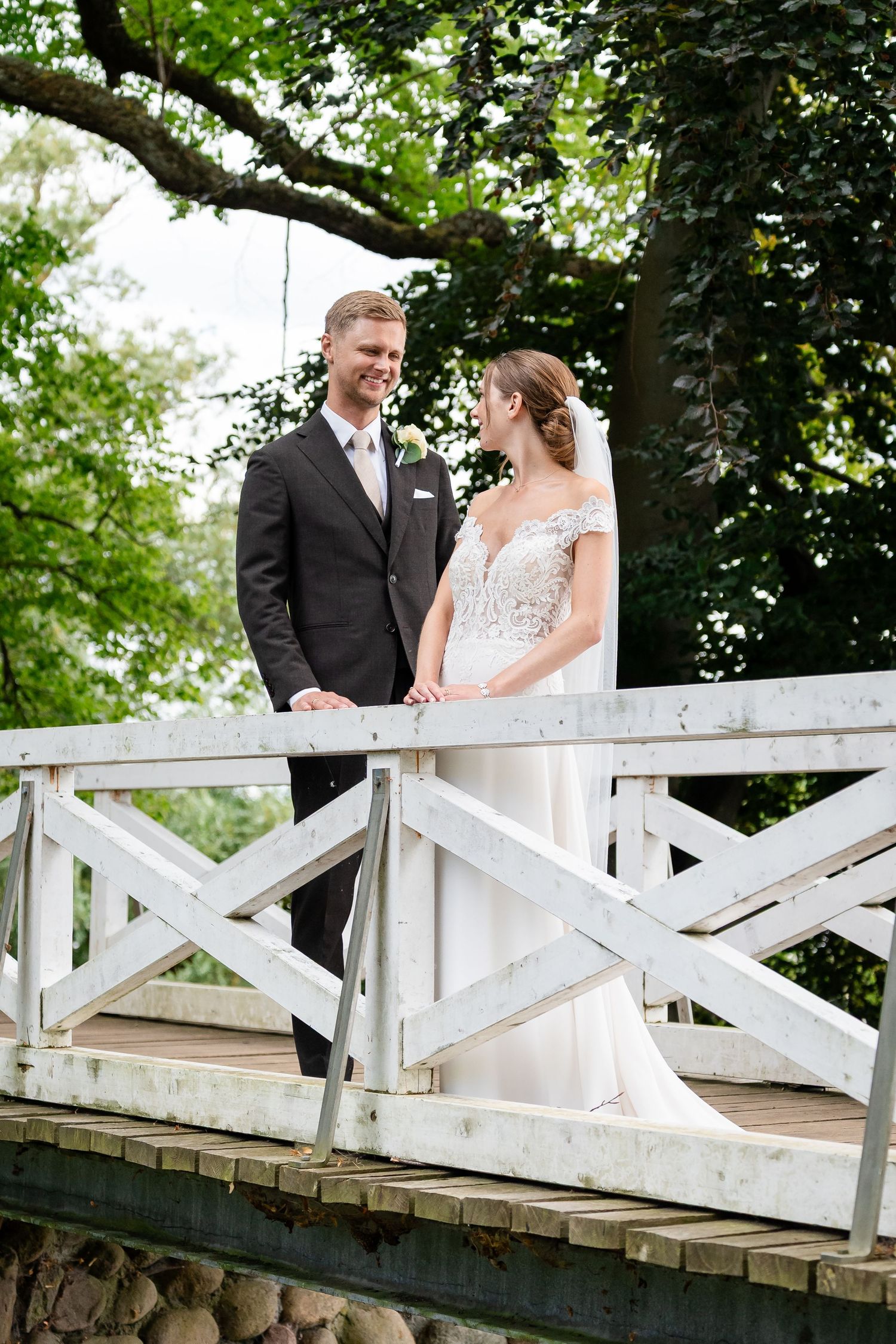 Bride and groom stand together on a white wooden bridge surrounded by lush green trees on their wedding day.