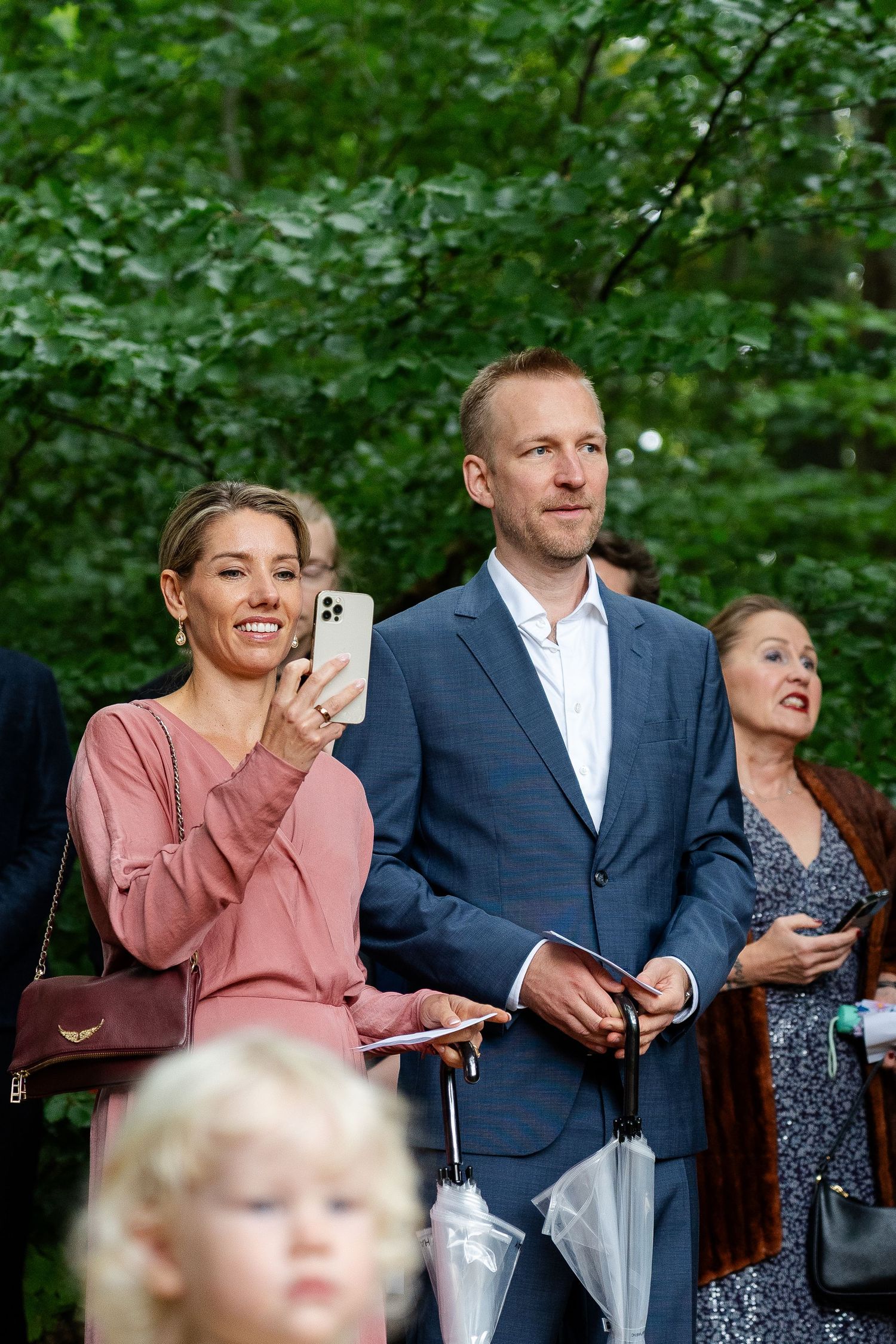 A smiling woman in pink takes a photo on her iPhone beside a man in a blue suit at an outdoor event.