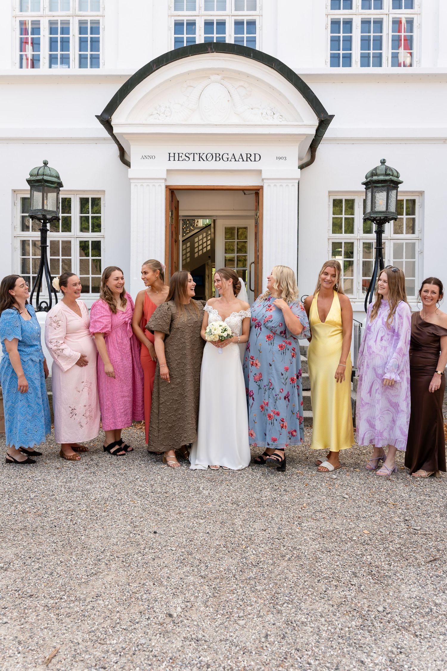 Bride in white gown poses with bridesmaids in colorful dresses outside Hestkøbgaard venue on a sunny day.