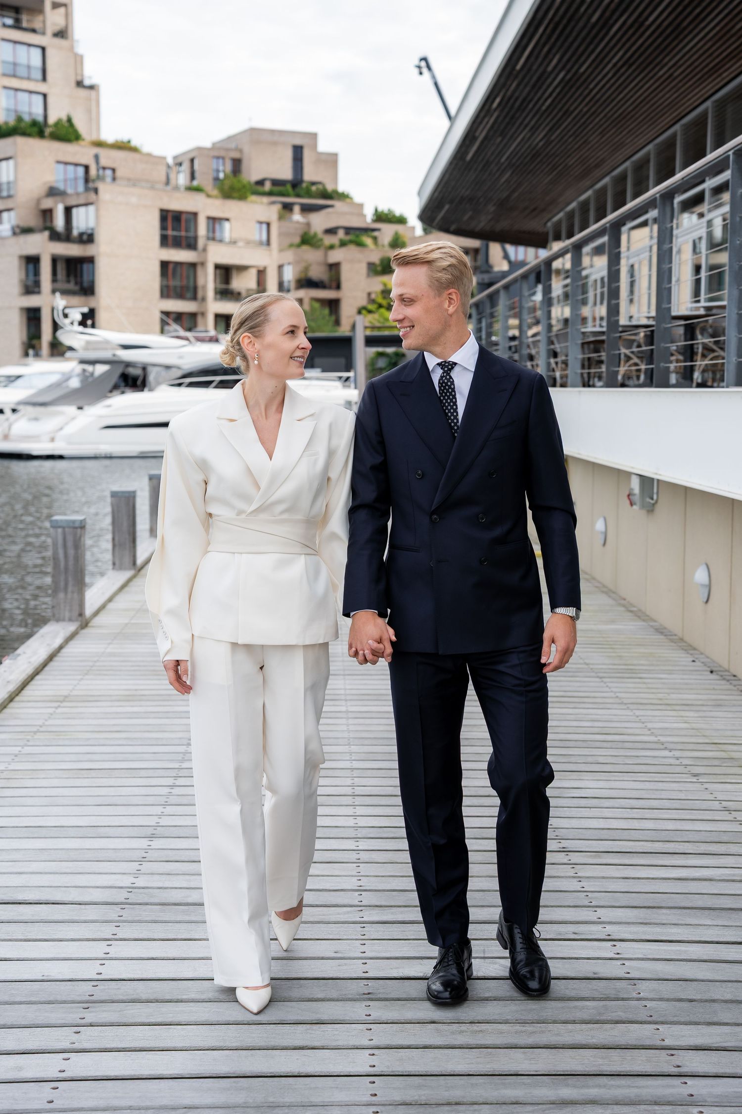 Elegant couple in matching white and navy suits holding hands on a marina boardwalk with yachts and modern buildings.