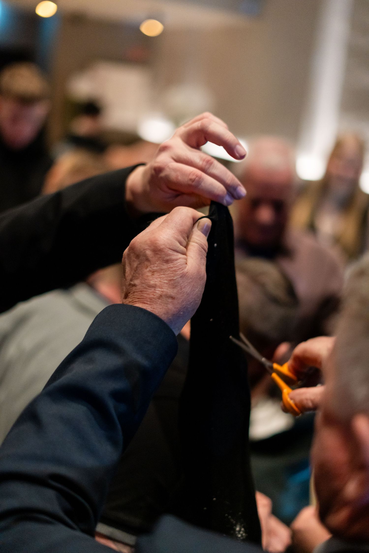 Hands using scissors to cut the foil cap off a wine bottle at a crowded indoor event.