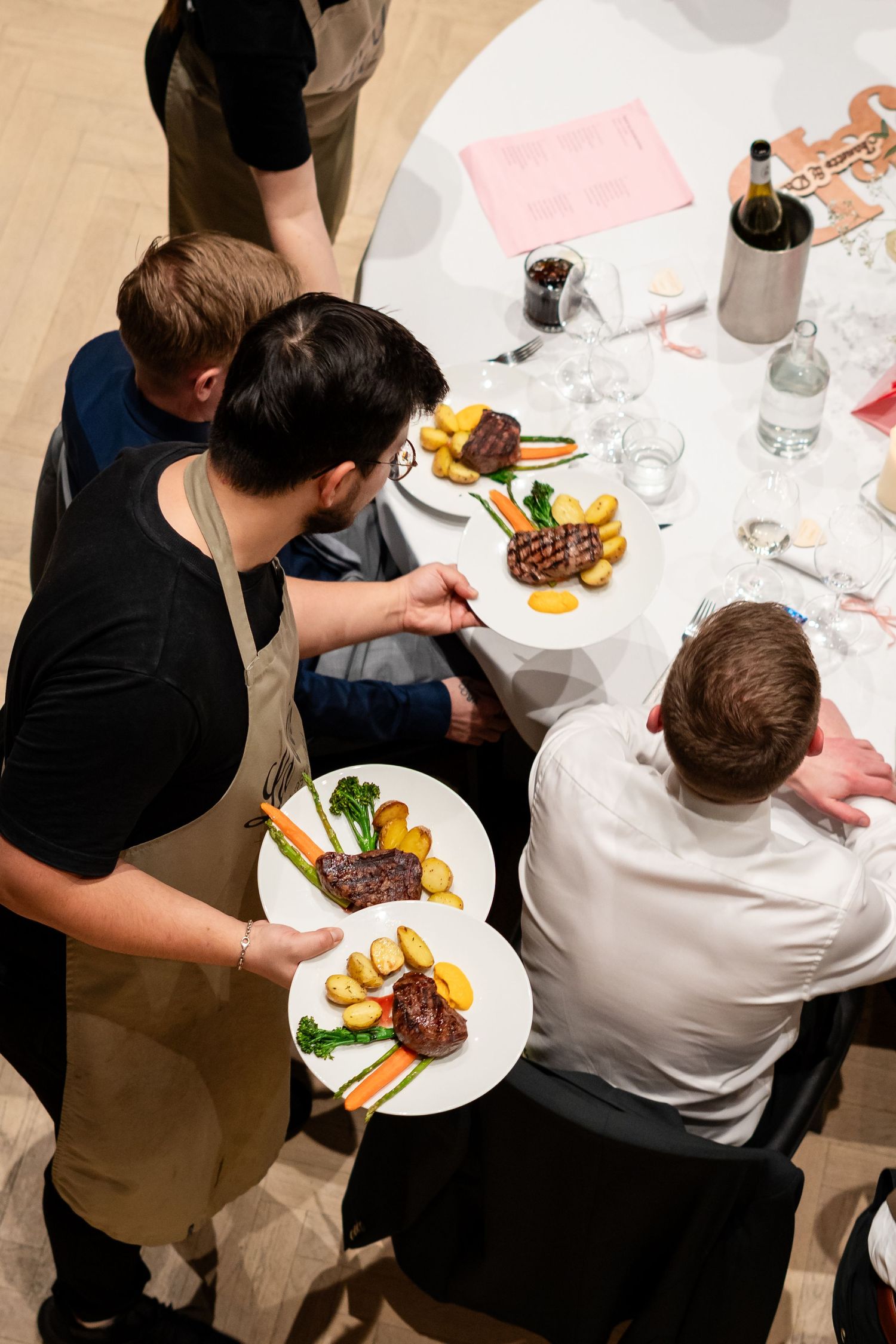 Overhead view of a chef serving plated steak dishes with roasted potatoes and vegetables at an elegant dining table.