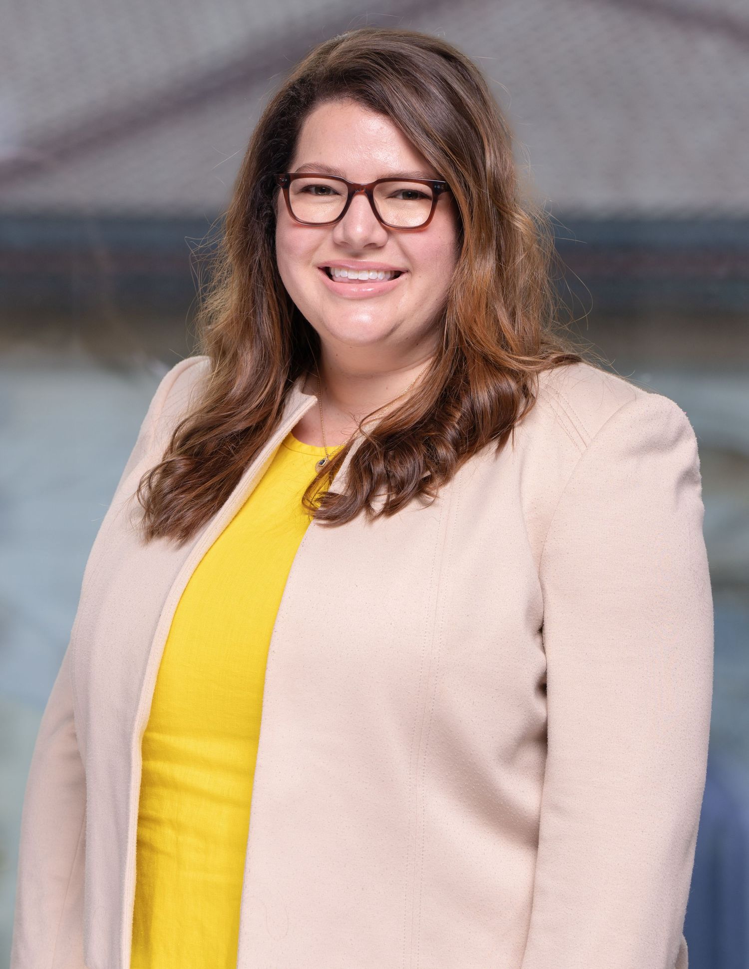 Professional headshot of person in glasses wearing a bright yellow top and beige blazer smiles warmly.