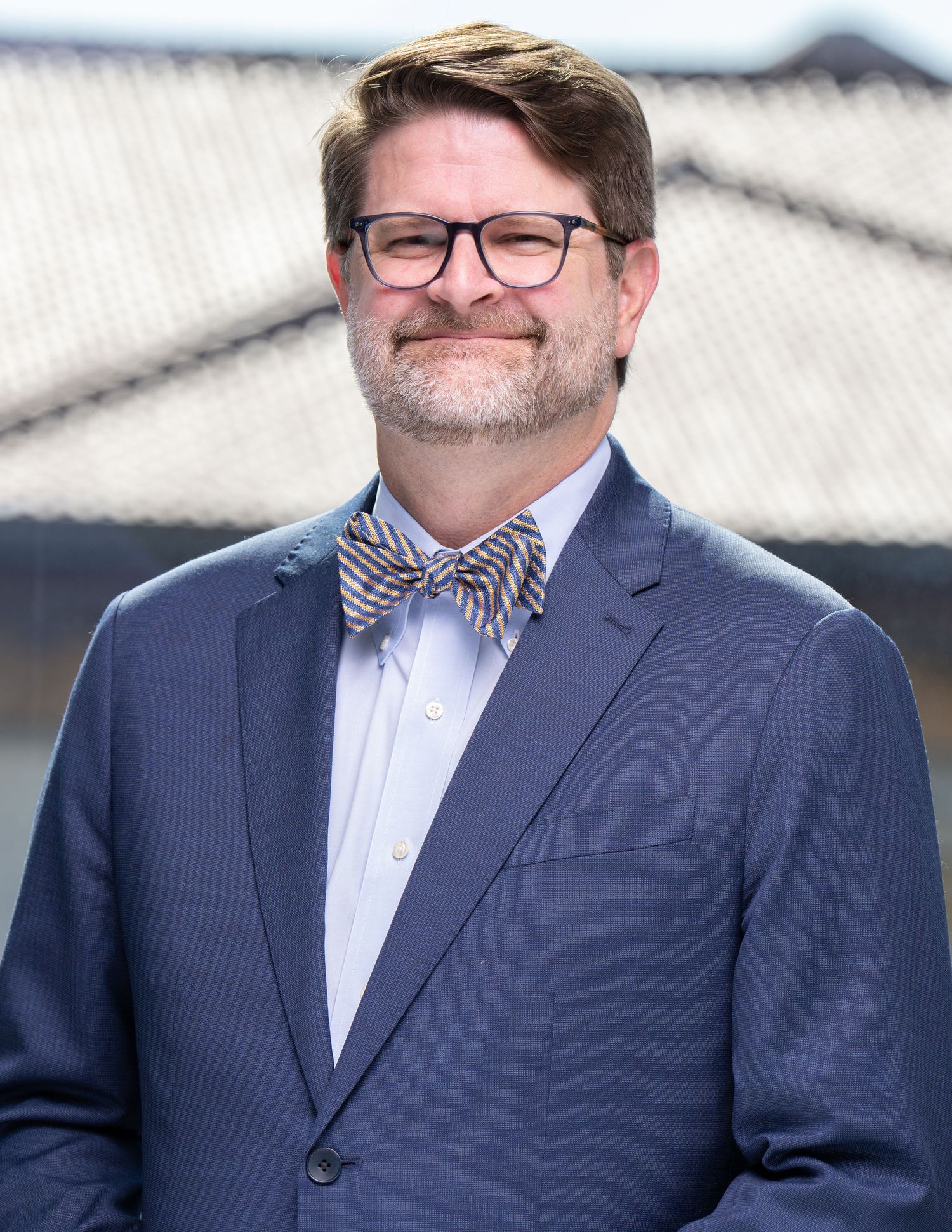 Professional headshot of person wearing a navy suit jacket, glasses, and bowtie smiles while looking to the side in a business portrait.