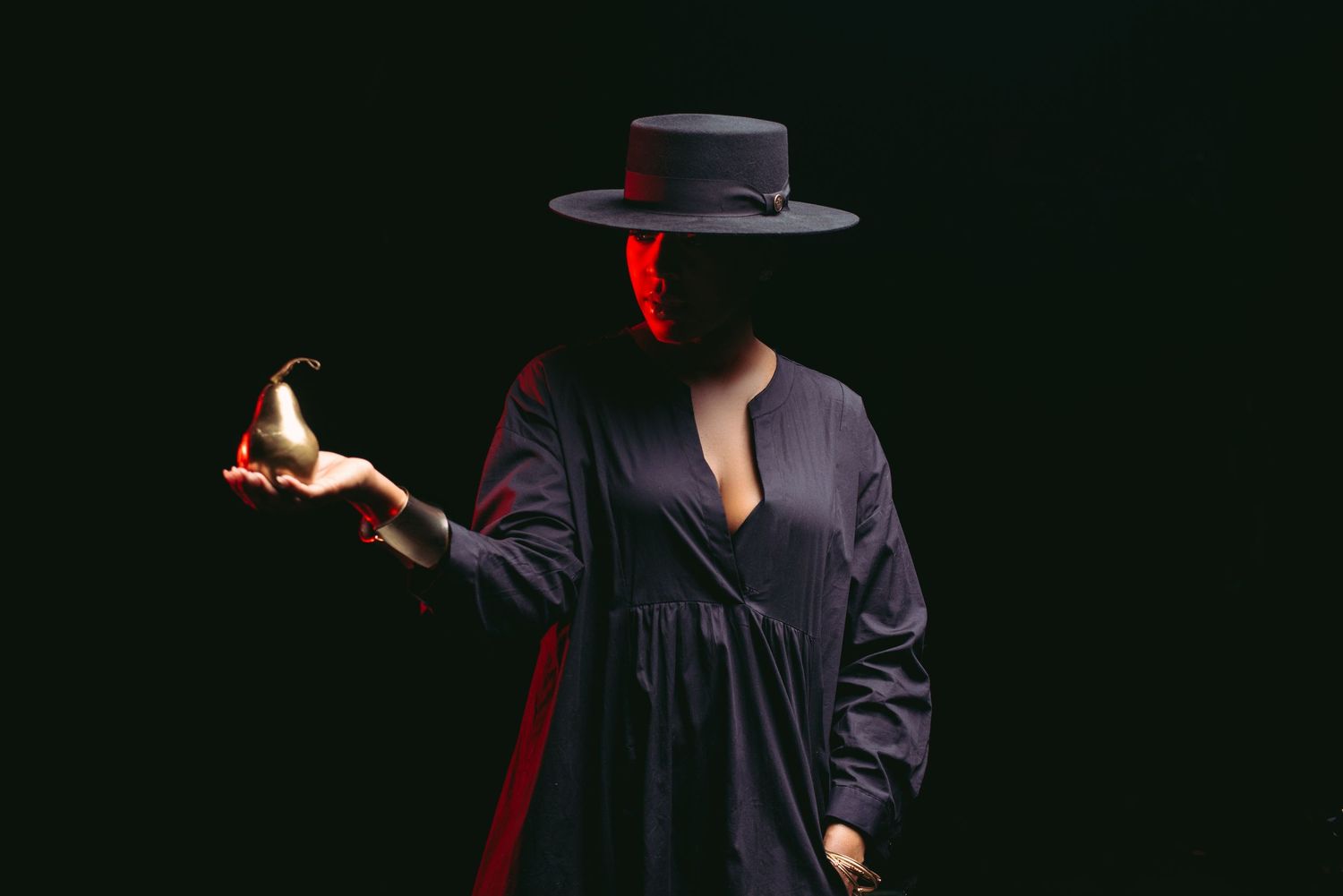 Mysterious figure in black dress and hat holds a flame against dark background.