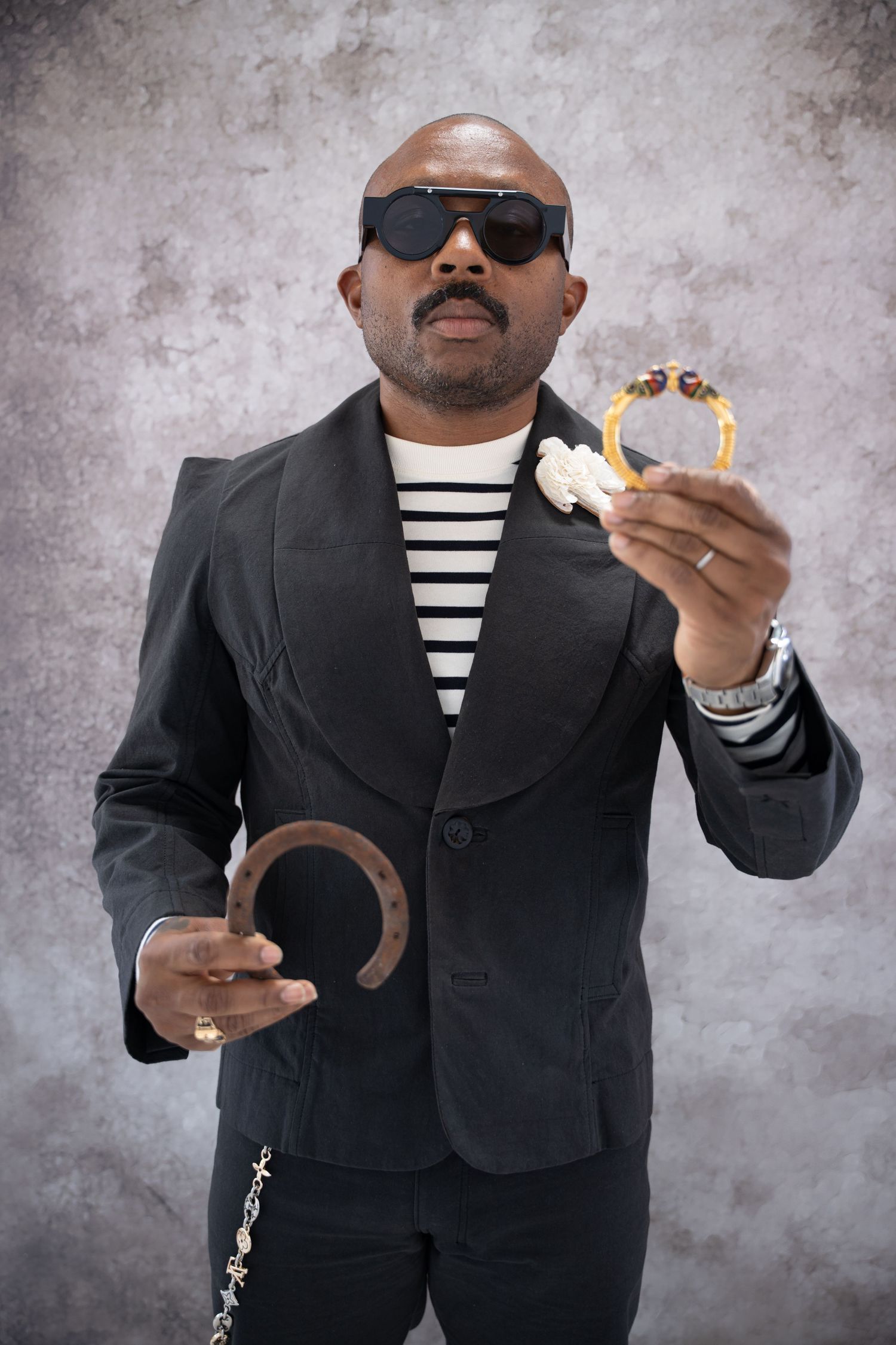 Person in black blazer and striped shirt holds a objects while posing against gray background for Labor Of Love series.