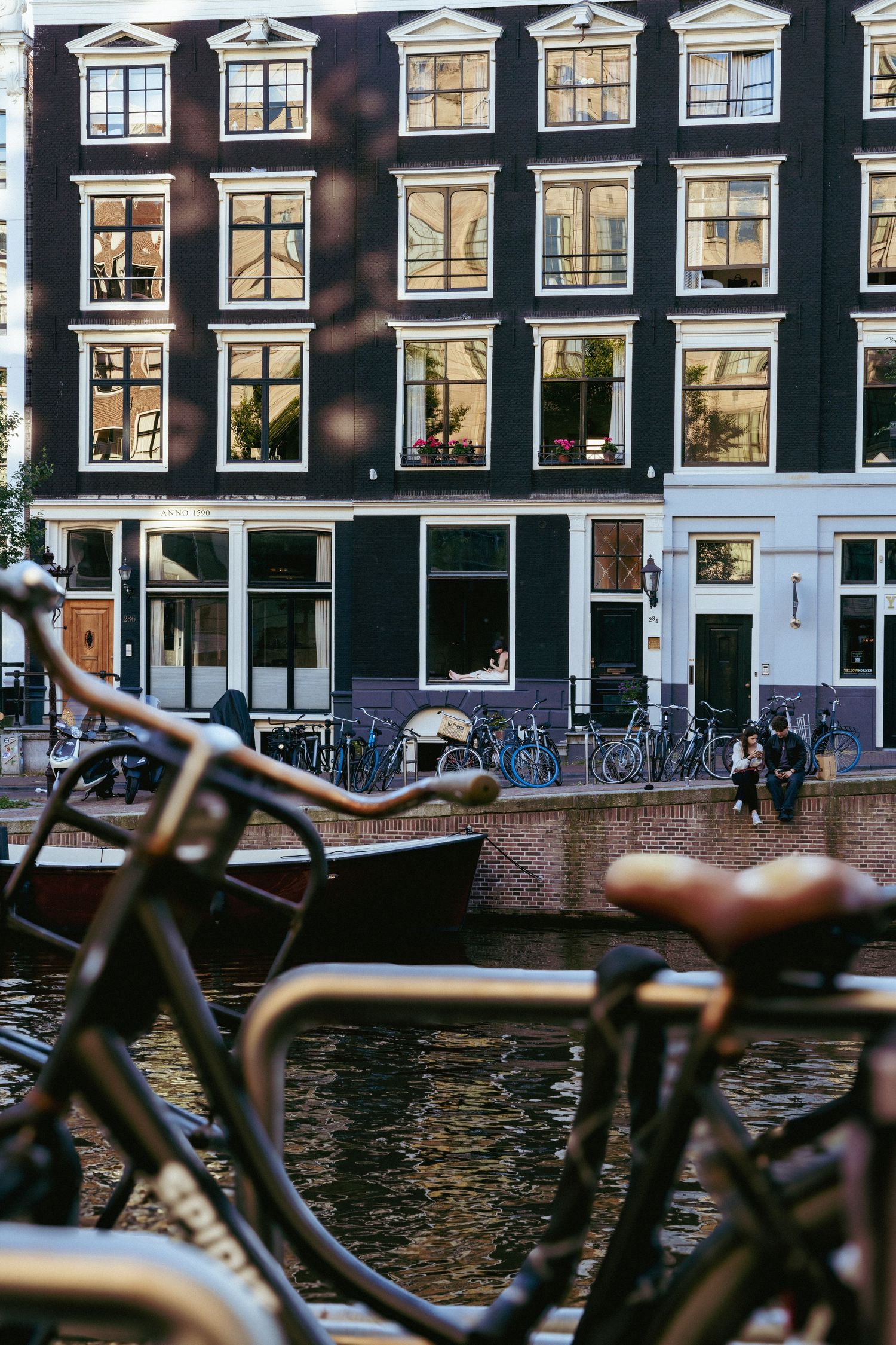 Traditional Amsterdam canal houses with tall windows reflect in the water as bicycles line the waterfront.
