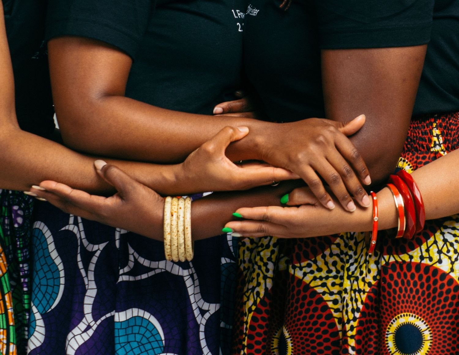 A row of people wearing colorful African print clothing and gold jewelry stand with their hands clasped together.