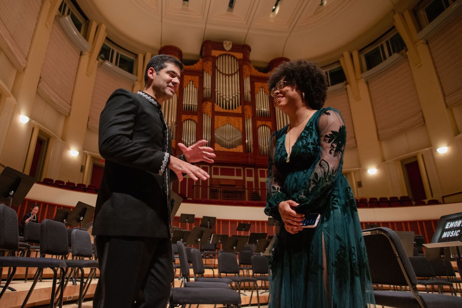 Concert performers on stage in formal attire with organ pipes visible in background.