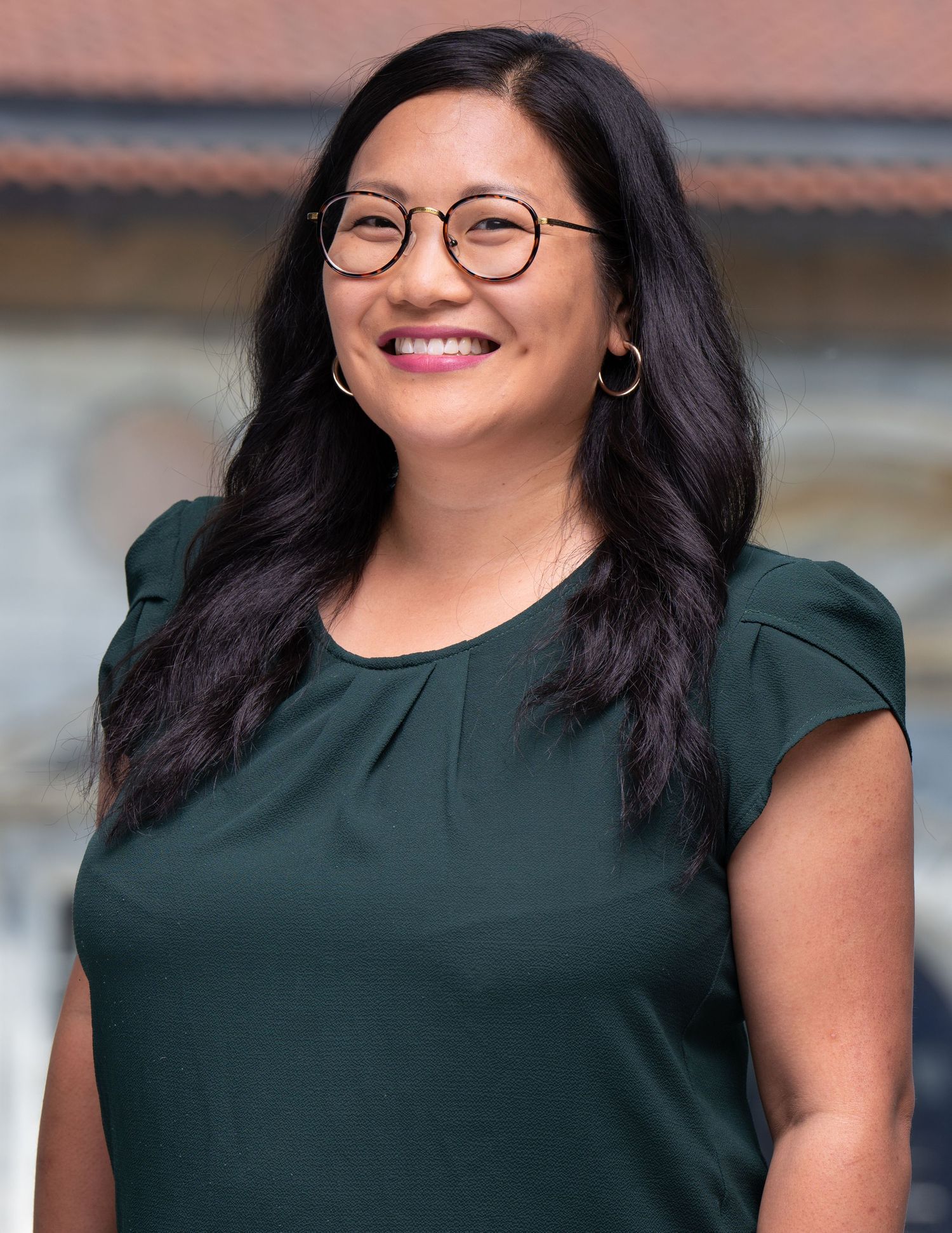 Professional headshot of a person in a dark green top wearing round glasses and silver hoop earrings against a blurred background.