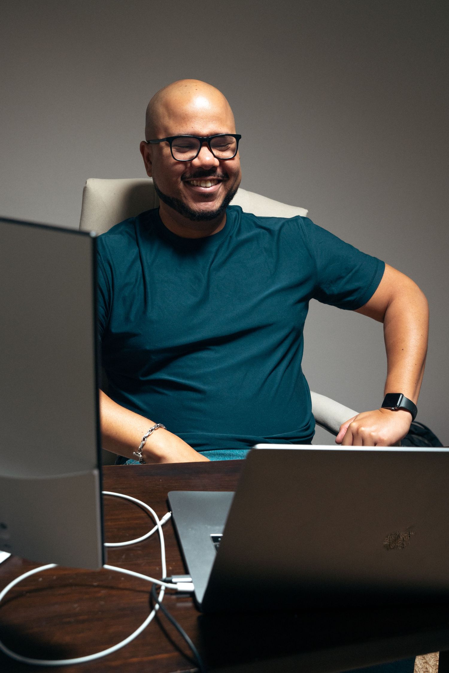Professional wearing glasses and teal shirt smiles while working at desk with laptop in office setting.