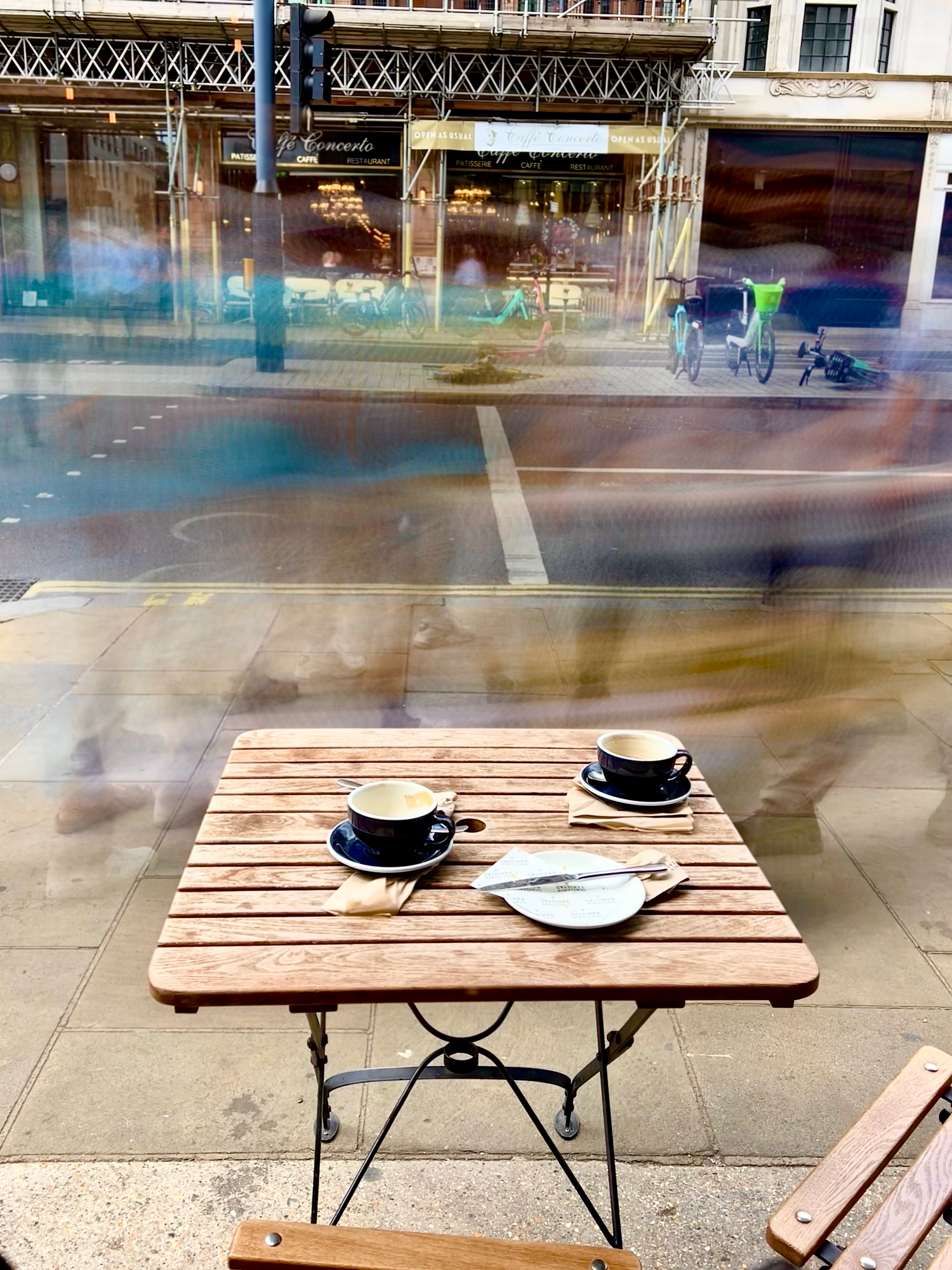 Two coffee cups on wooden table at outdoor cafe with blurred urban street scene in background.