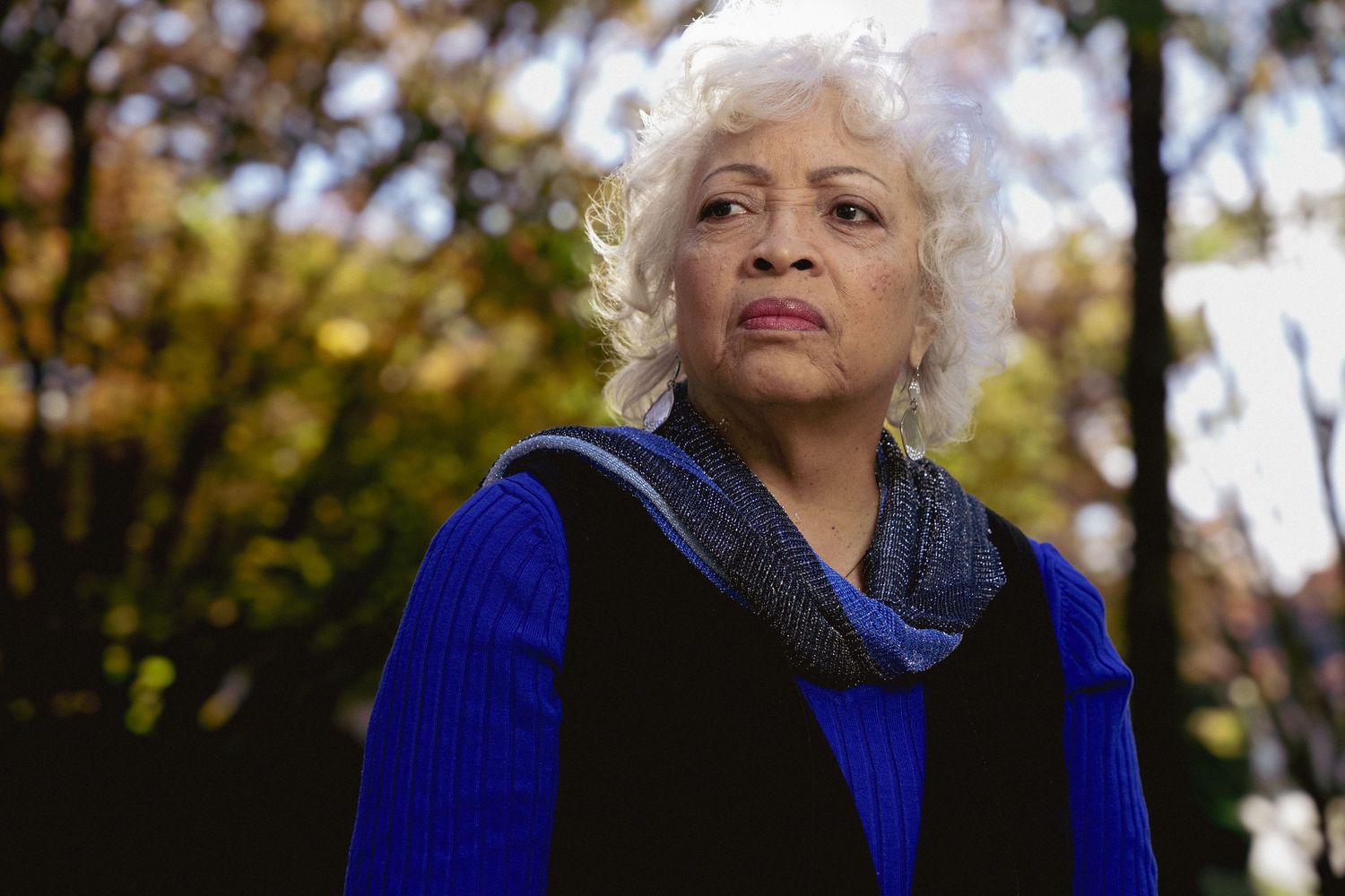 Elderly woman with white curly hair wearing a blue top and gray scarf, standing outdoors with autumn trees in background.