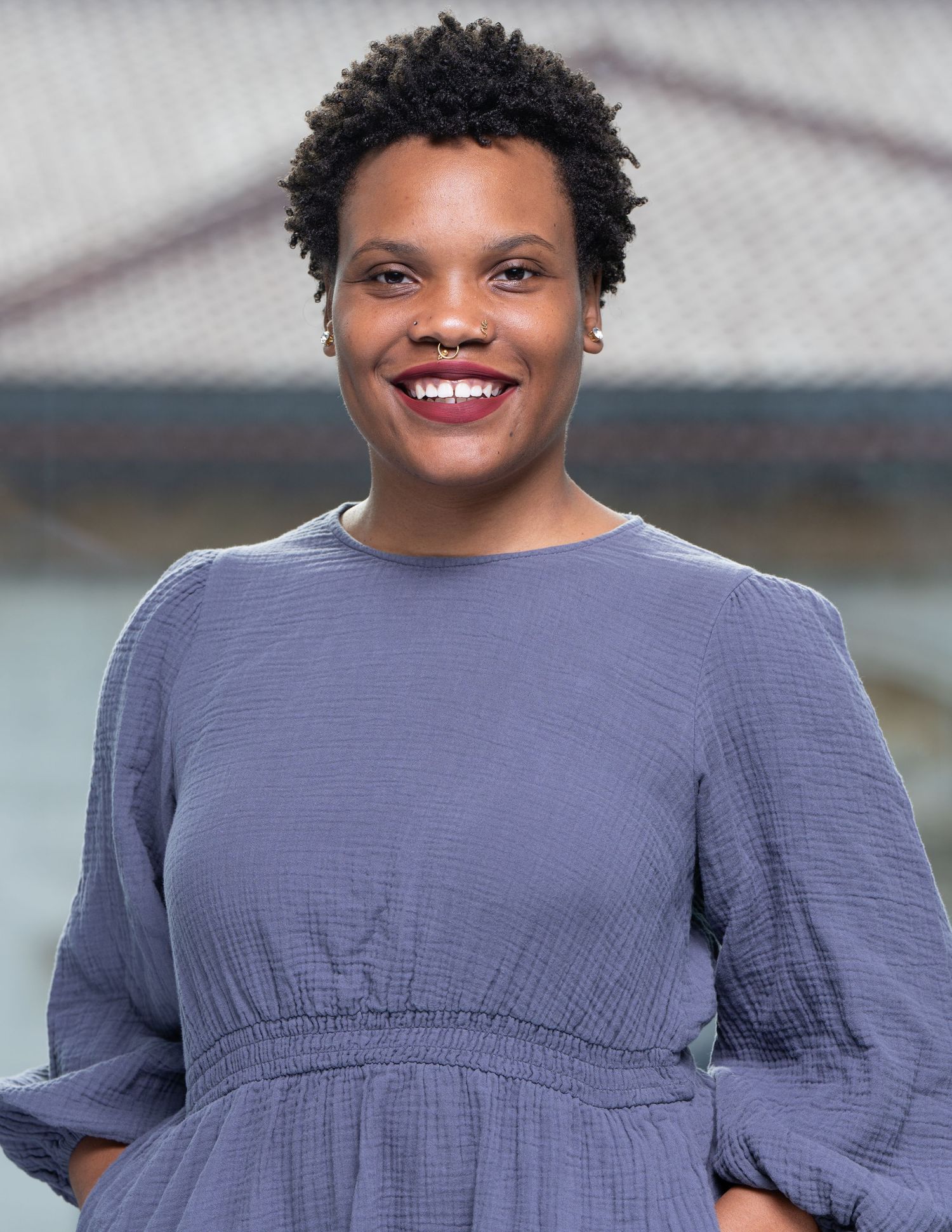 Professional headshot of woman wearing a blue-gray dress smiling against a blurred outdoor background.