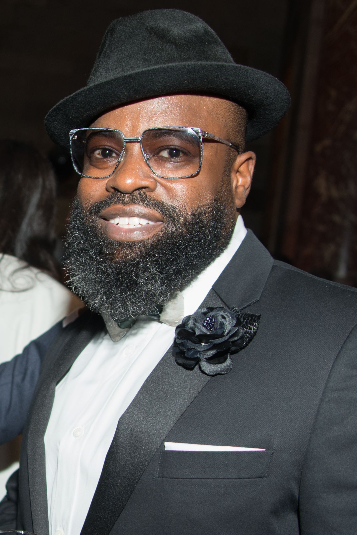 Stylish man in black suit and hat with full beard wearing glasses and smiling at formal event.