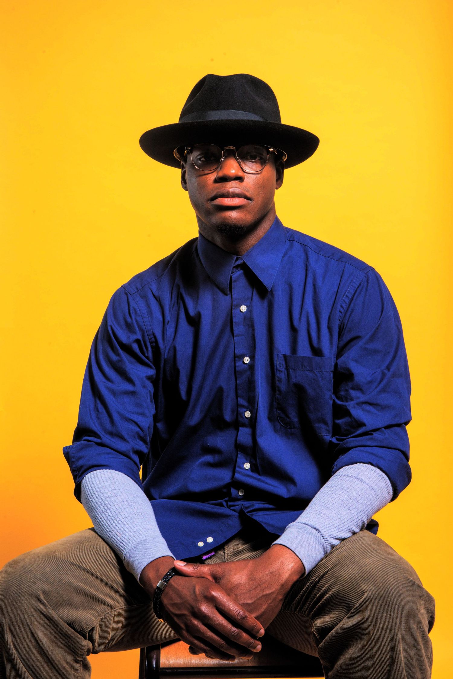Portrait of person in blue shirt and black hat seated against vibrant yellow background.