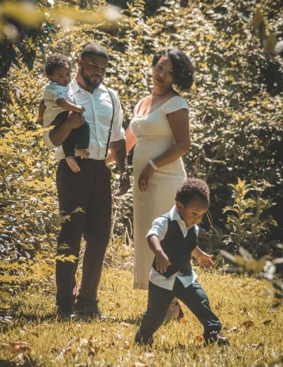 A joyful Black family poses outdoors on green grass, with a pregnant mother, father holding a toddler, and young son running.
