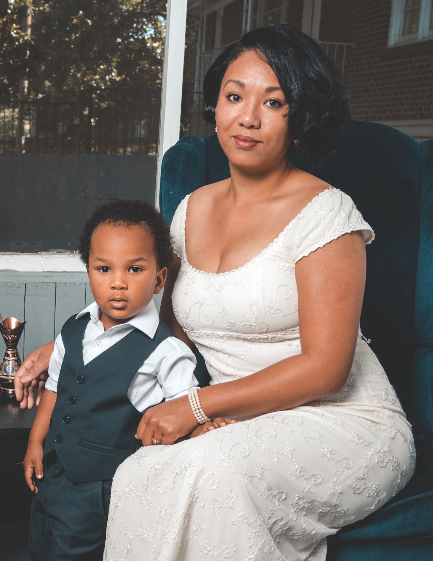 A bride in a white lace dress sits with a young child in formal attire during an outdoor photo session.