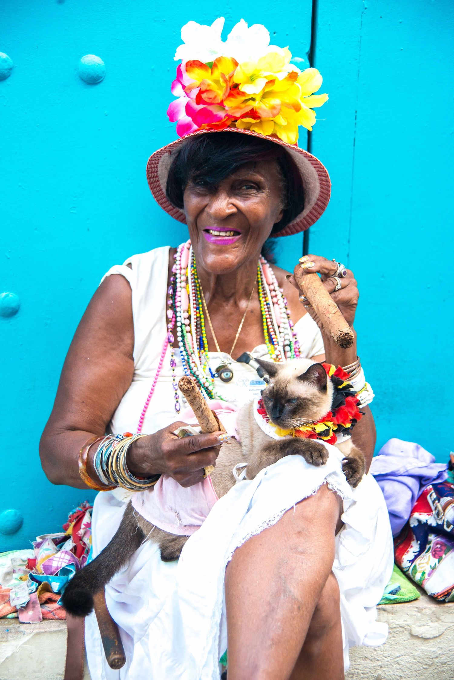 A woman wearing a sun hat and white dress holds a small dog while sitting against a bright blue wall.