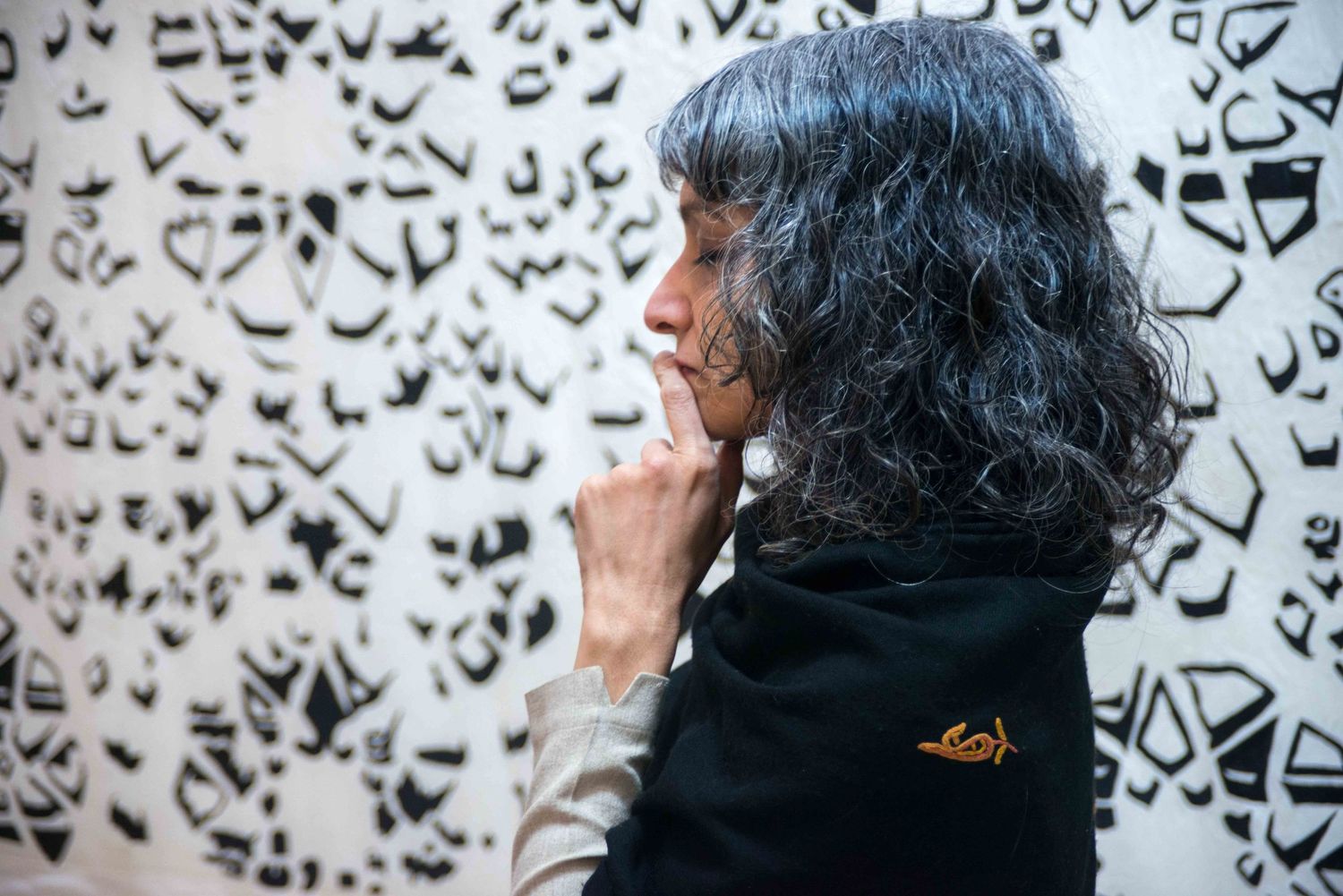 A person with curly dark hair contemplates against a patterned wall art background.