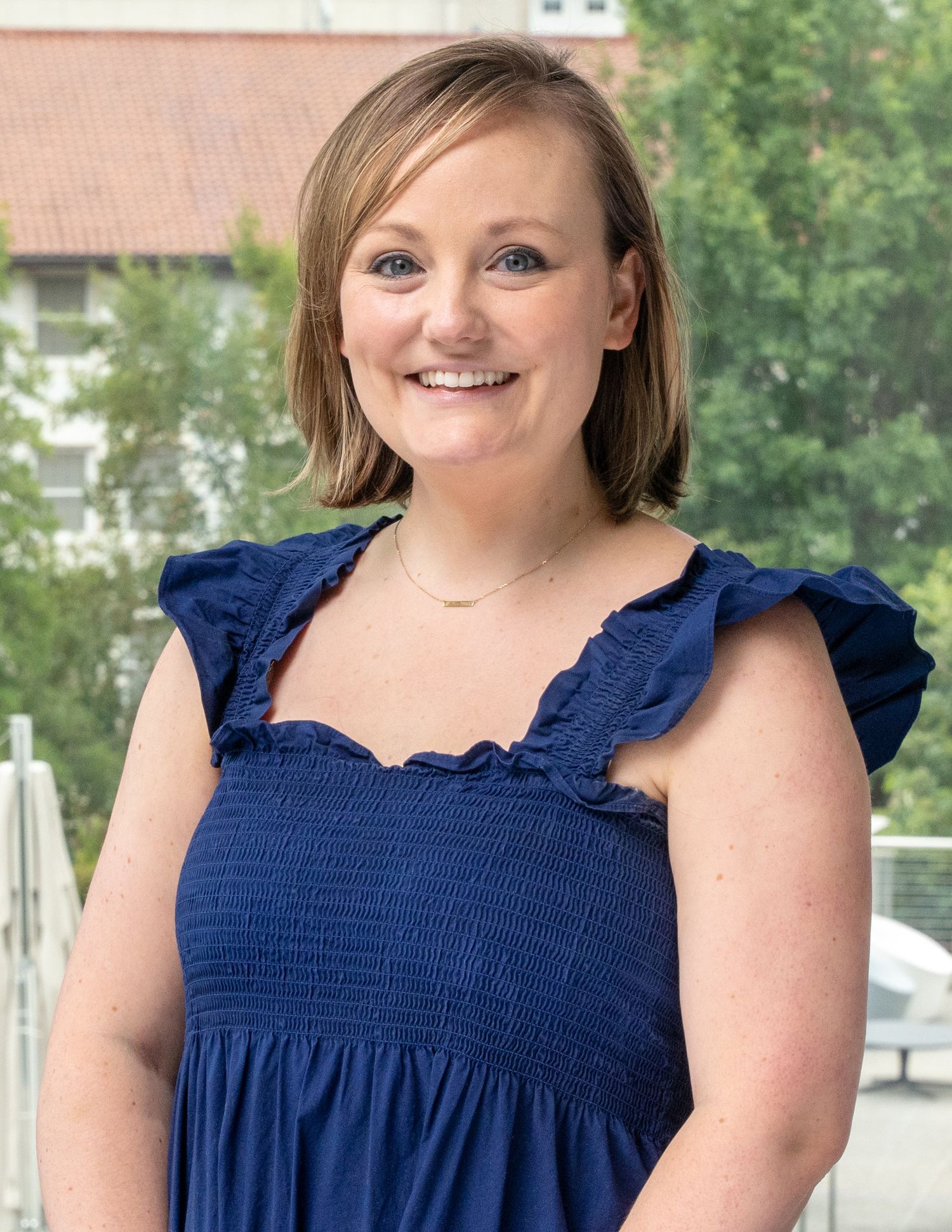 Professional headshot of woman in navy blue smocked dress smiling outdoors with buildings and trees in background.