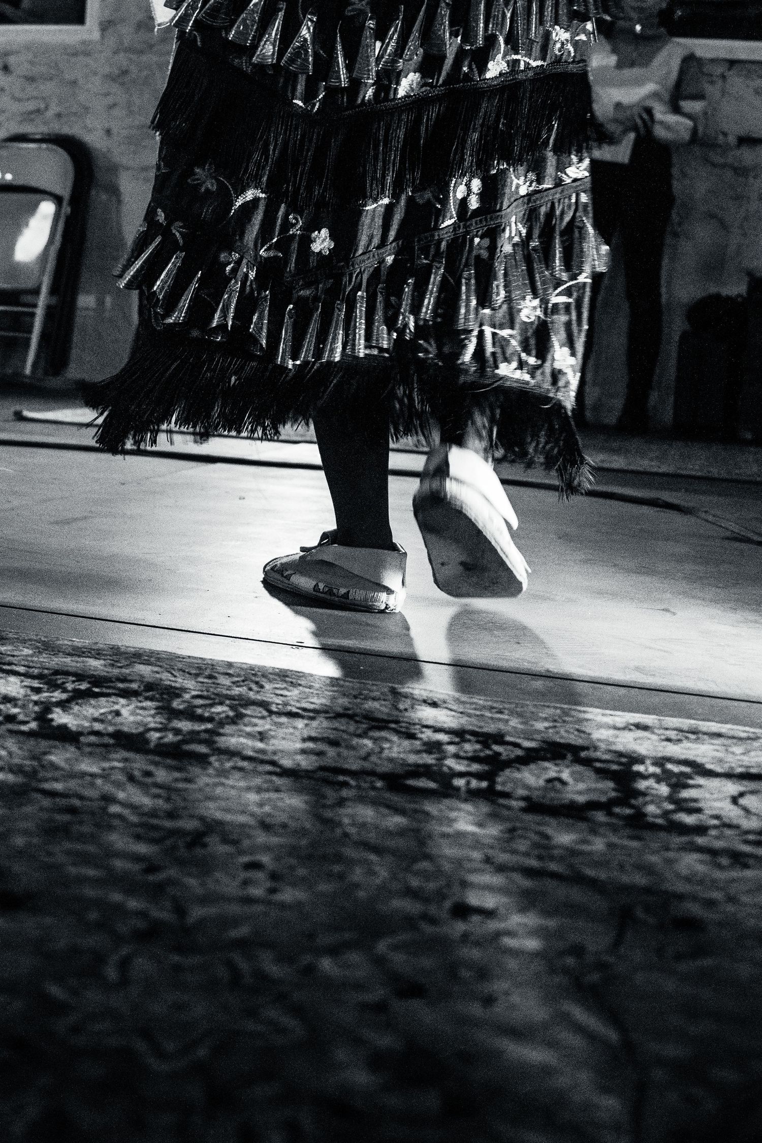 Black and white photo of fringed garments and feet on a textured floor.