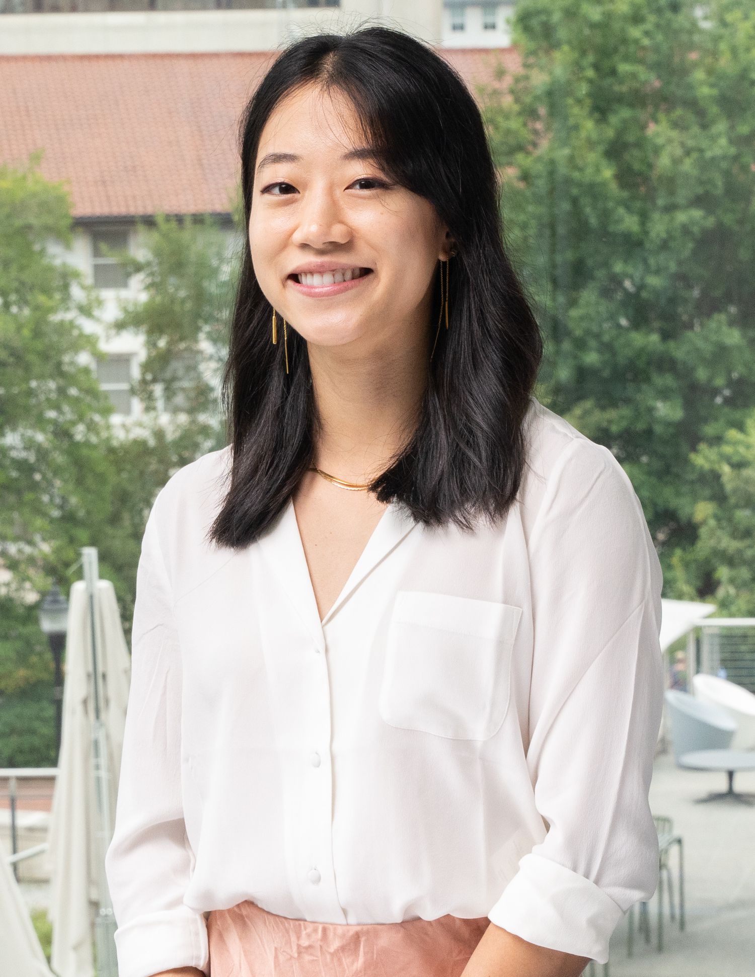 Professional headshot of person in a white shirt and pink apron smiles warmly while standing outdoors near buildings and trees.