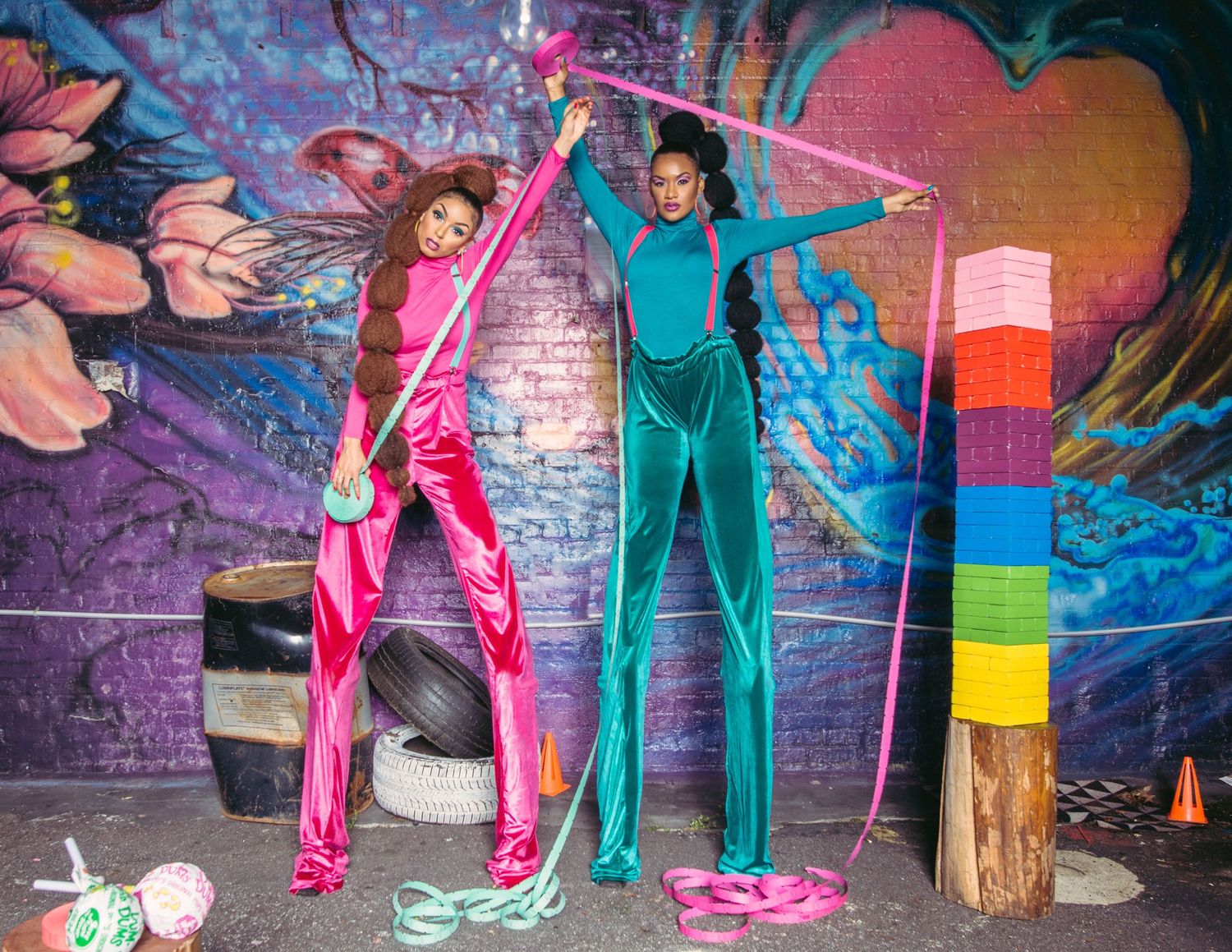 People in pink and teal tracksuits perform dance moves in front of a graffiti wall at night.