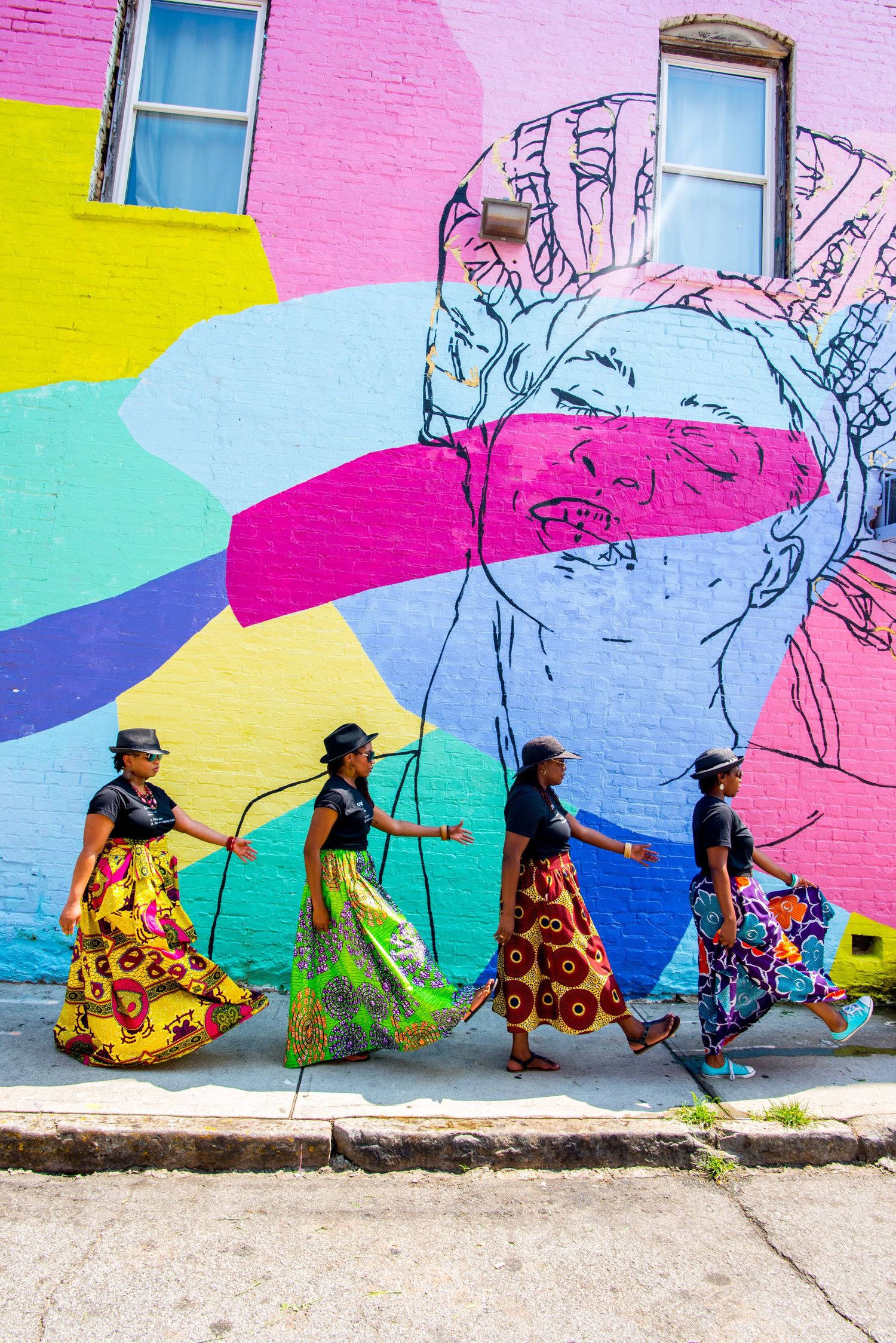 Colorful street art mural serves as backdrop for people walking past in vibrant floral dresses and hats.
