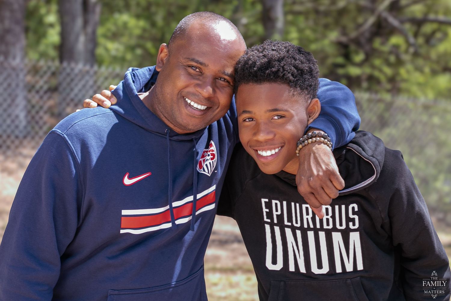 Two smiling men pose together outdoors, one wearing a USA hoodie and the other an E Pluribus Unum sweatshirt.