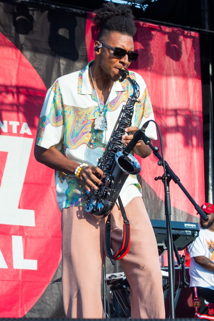 Musician performs on stage with a saxophone wearing a colorful Hawaiian shirt and khaki pants against a red backdrop.