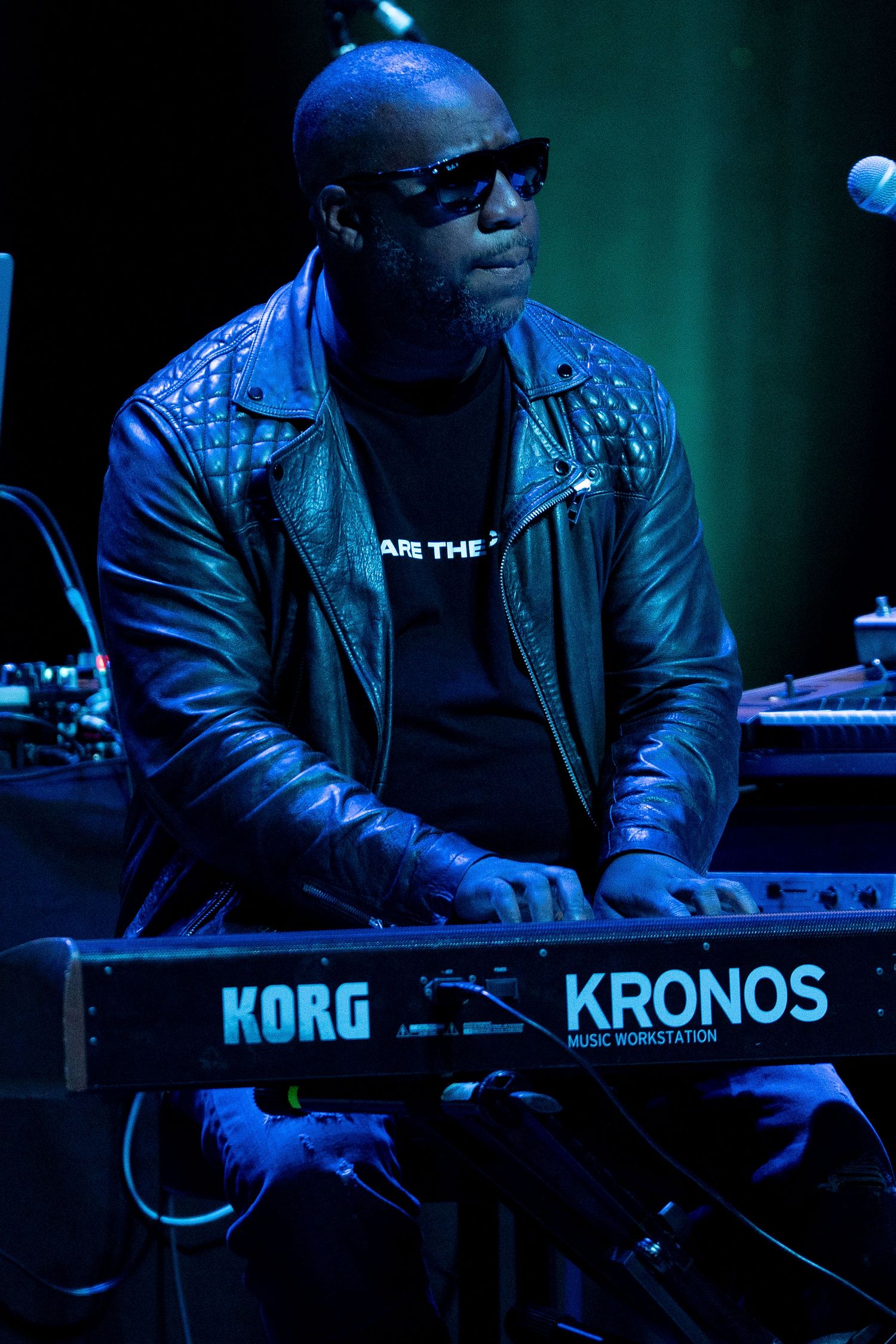 Musician in leather jacket performing on a Korg Kronos keyboard in dark lighting.